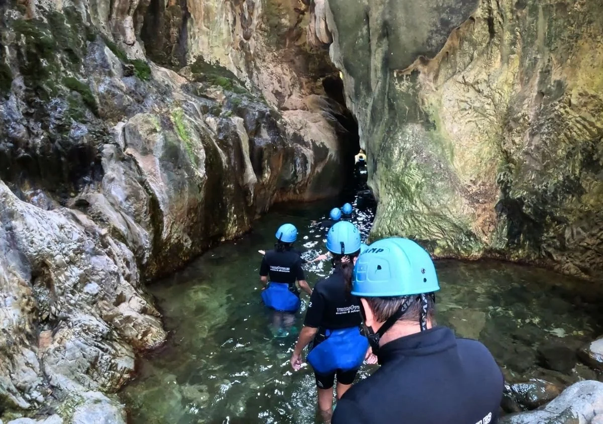 A group of people wearing blue helmets and canyoning gear wade through clear water in a narrow, rocky gorge along the Guadalmina River.