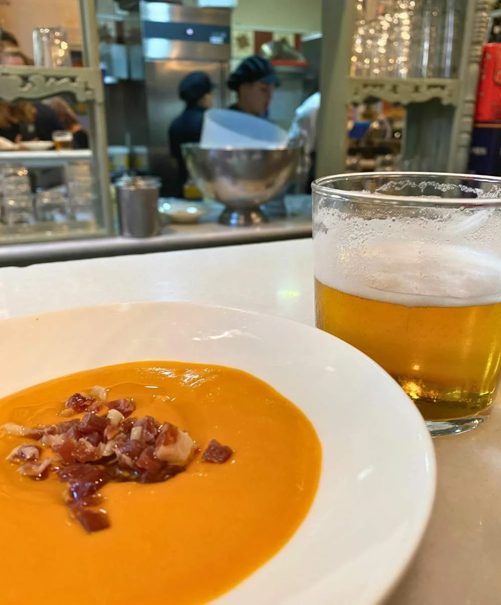 Bowl of creamy orange soup topped with diced cured ham on a white plate beside a glass of golden beer, with a softly blurred open kitchen and restaurant interior in the background.