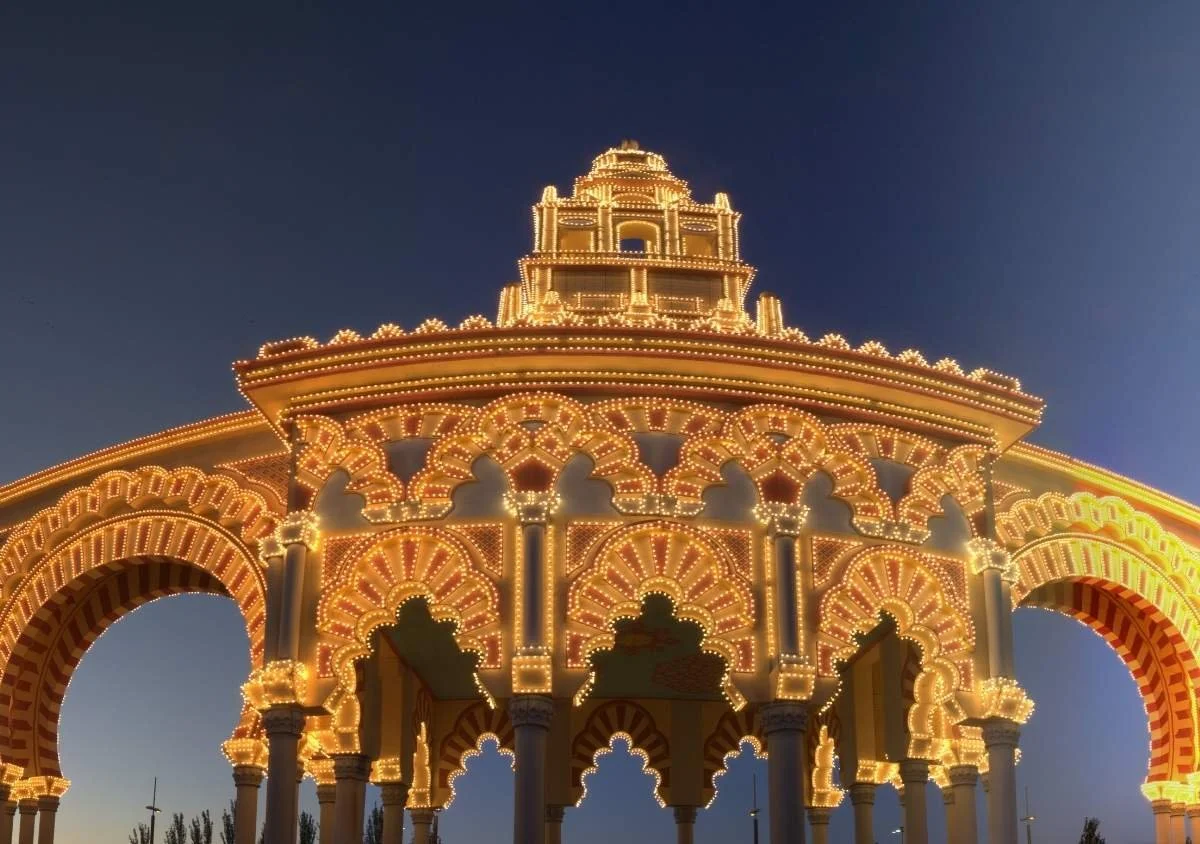Illuminated ornate pavilion at dusk, featuring Moorish-style arches and intricate decorative patterns outlined with warm golden lights against a deep blue evening sky.