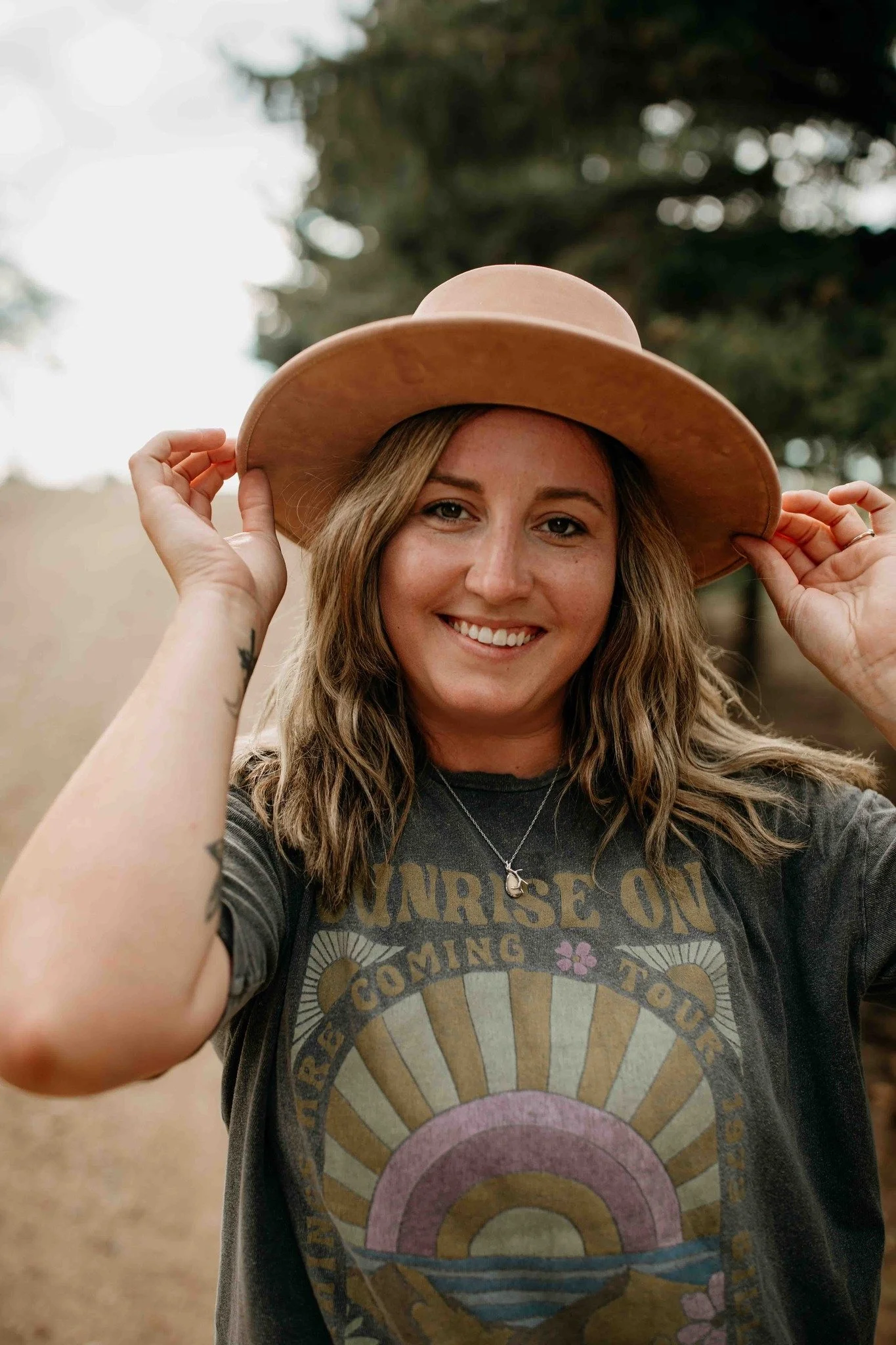 A woman with long wavy hair holding a wide-brimmed tan hat on her head, smiling outdoors with trees blurred in the background.