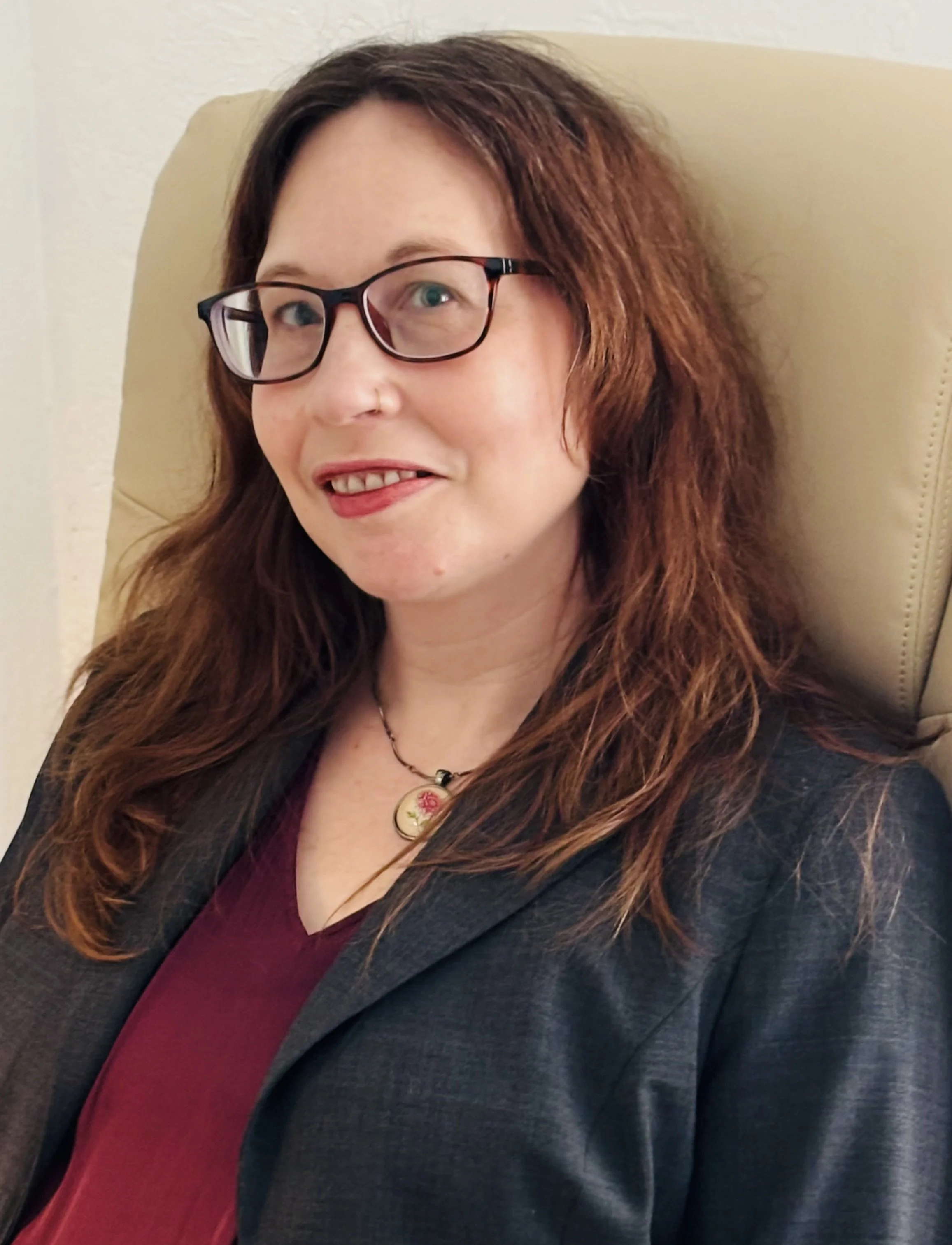 A woman with long, wavy, reddish-brown hair and glasses, wearing a dark blazer and a maroon top, sitting on a beige chair against a neutral wall.