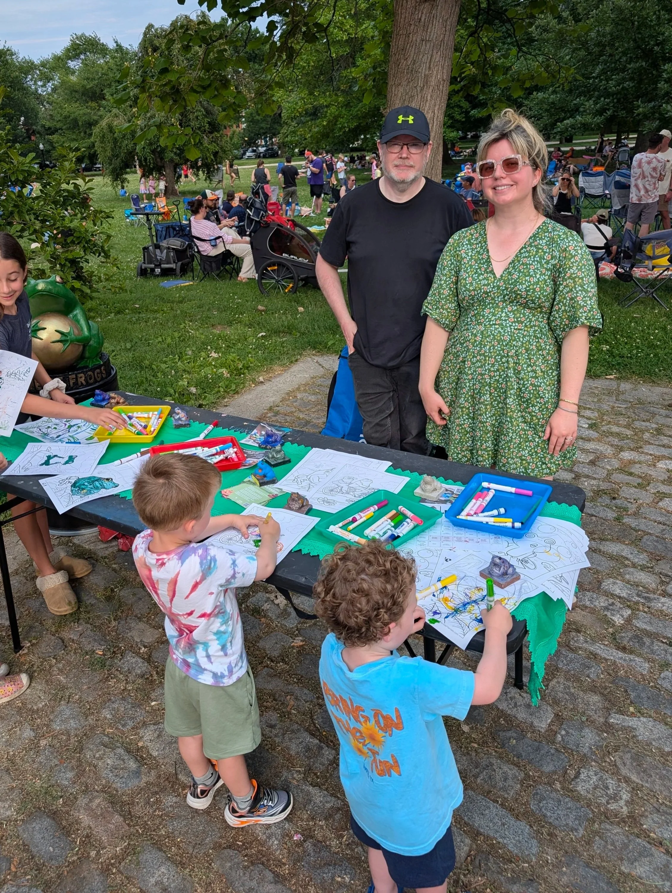 People at an outdoor park event. Two adults are posing for a photo, children are drawing and coloring at a table with art supplies. The crowd is gathered with chairs and blankets under trees, enjoying a sunny day.