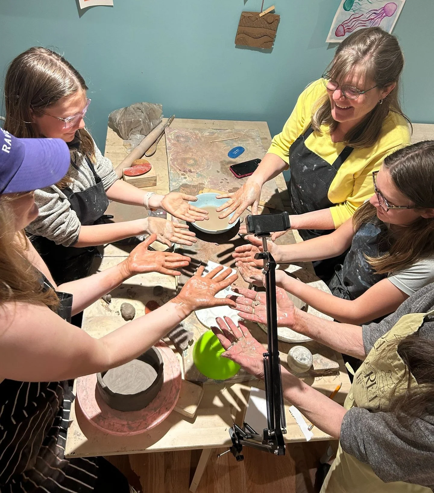 A group of people at a pottery workshop, working with clay on a worktable, surrounded by pottery tools, with some people using a smartphone to record or take pictures.