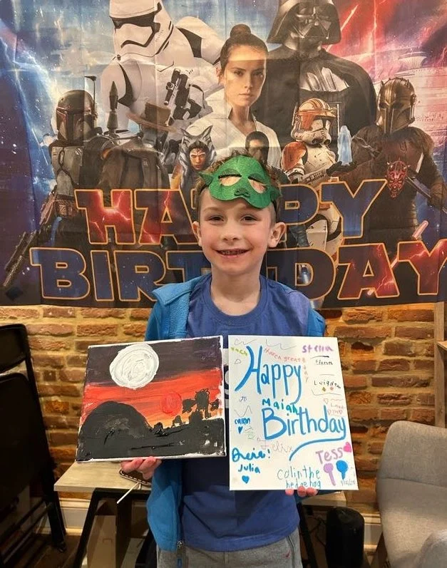 A young boy wearing a green mask costume and a blue shirt, holding a drawing and a birthday card at a Star Wars themed birthday party. The background features a large poster with Star Wars characters and a 'Happy Birthday' message.