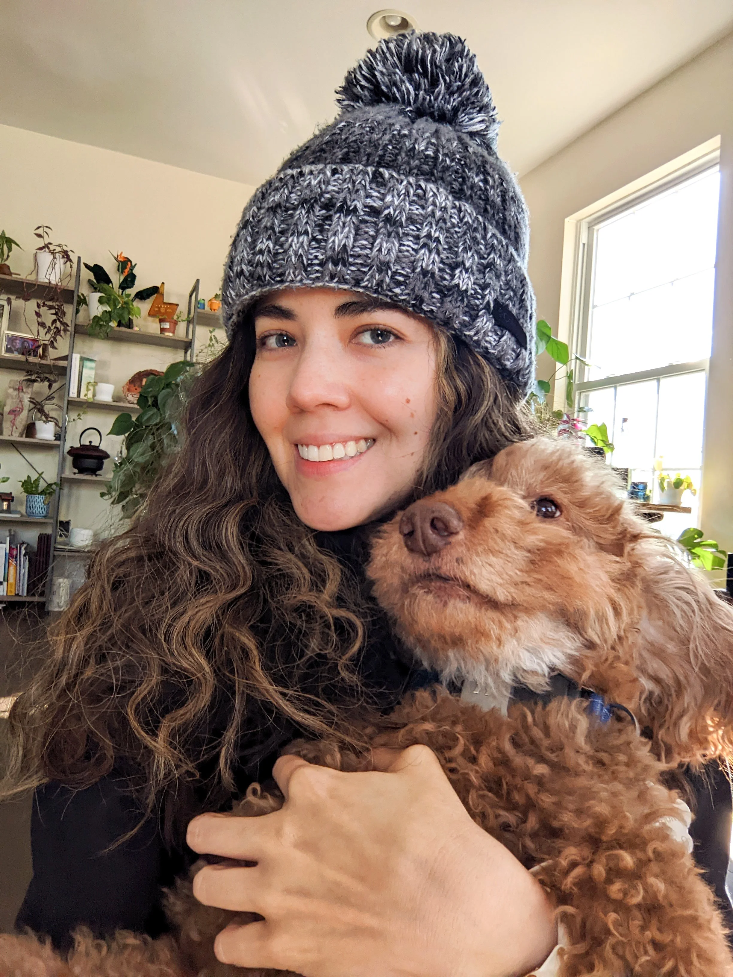 A young woman with long curly hair wearing a gray knit beanie with a pom-pom, smiling and holding a small brown dog, near a window with plants and shelves in the background.
