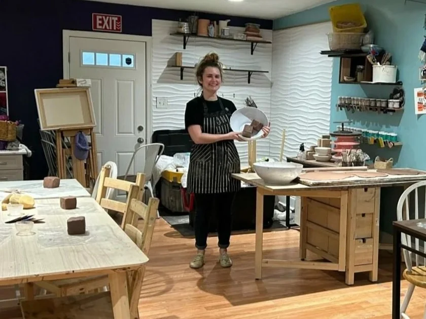 Young woman in black apron holding a block of clay, smiling in pottery or craft studio with tables, shelves, and art supplies.