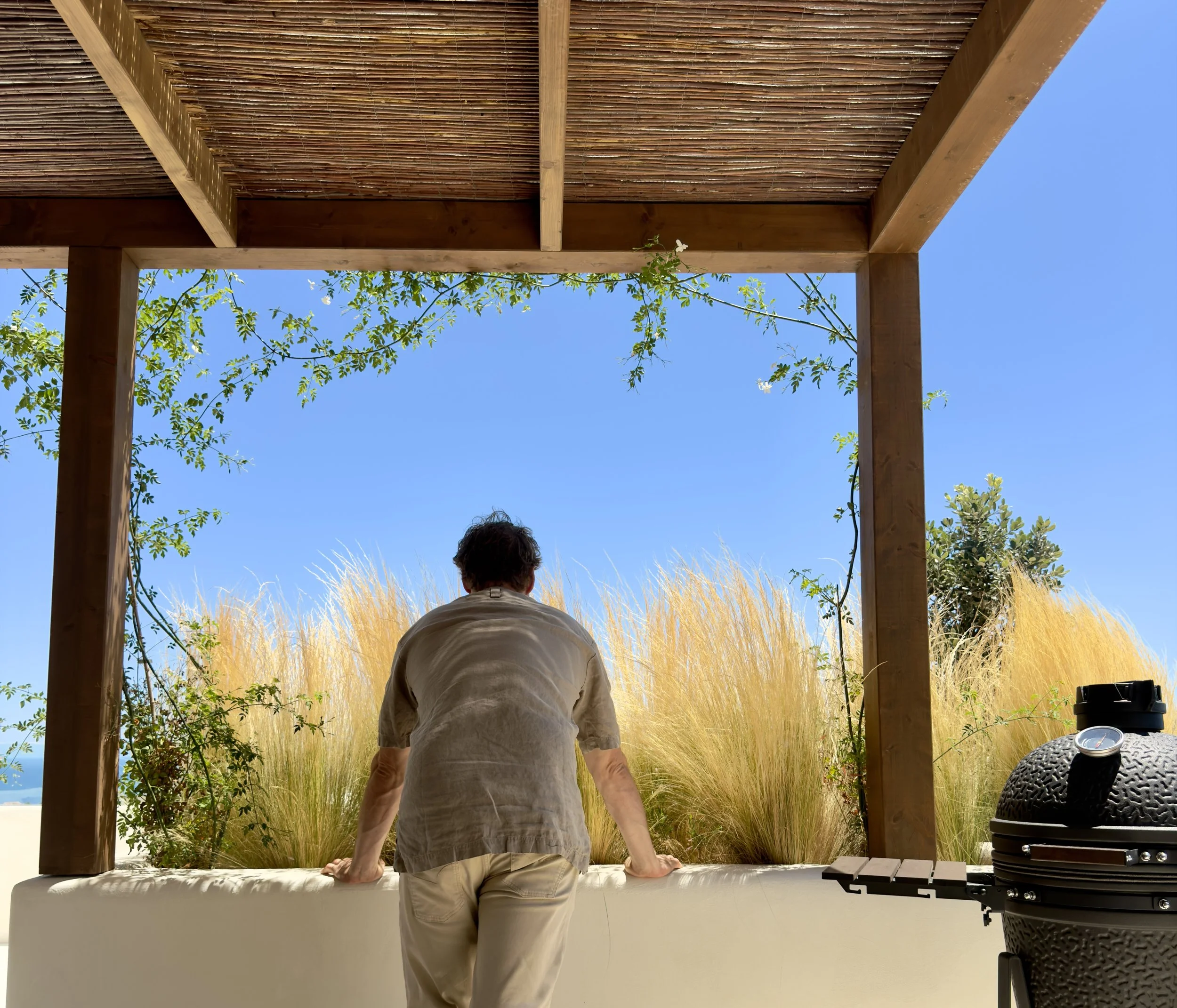 A person sitting on a ledge under a wooden pergola with a woven roof, overlooking tall grass and trees, with a clear blue sky in the background.