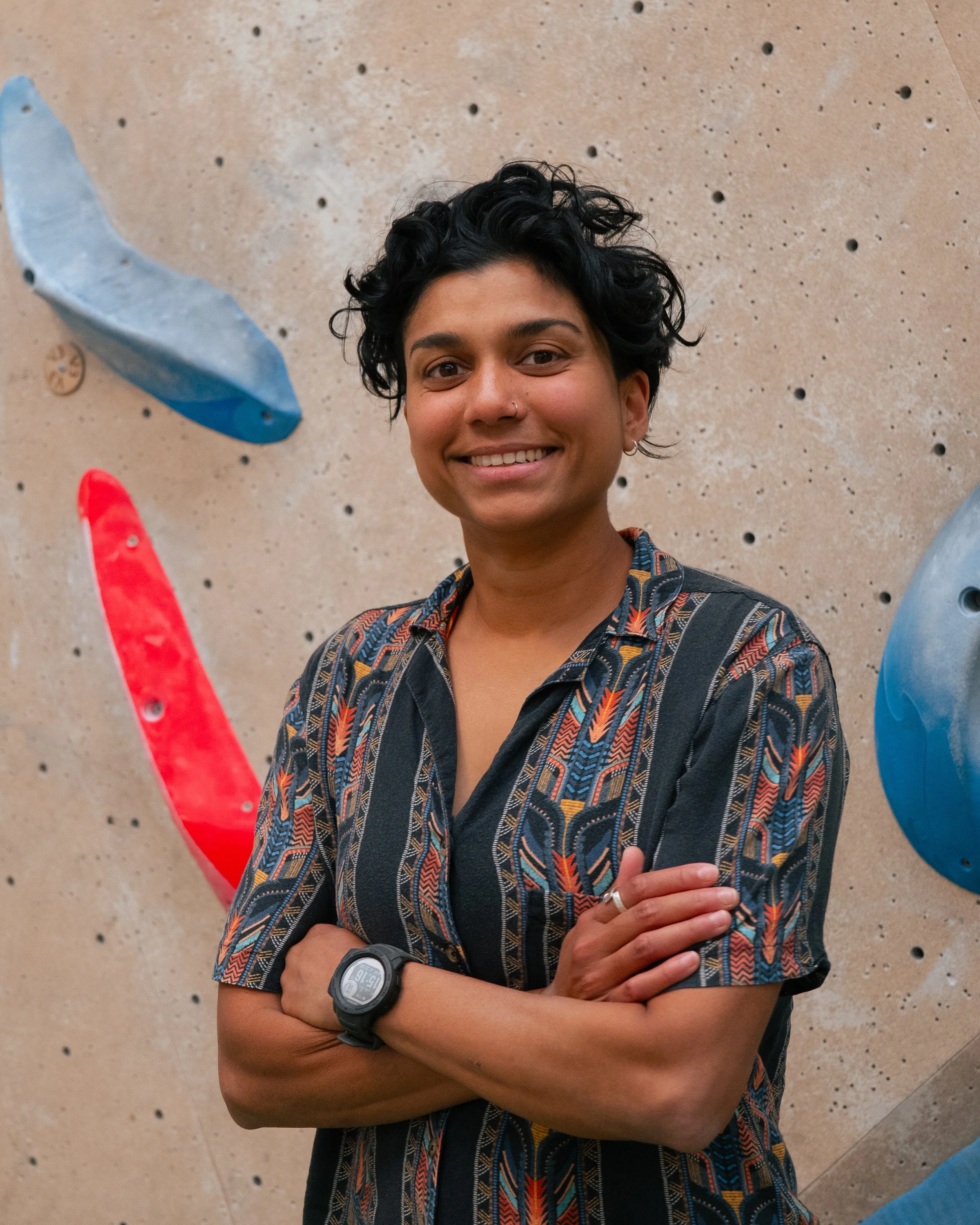 A person with dark, curly hair and a nose ring, smiling with arms crossed, standing against a textured climbing wall with colored holds.