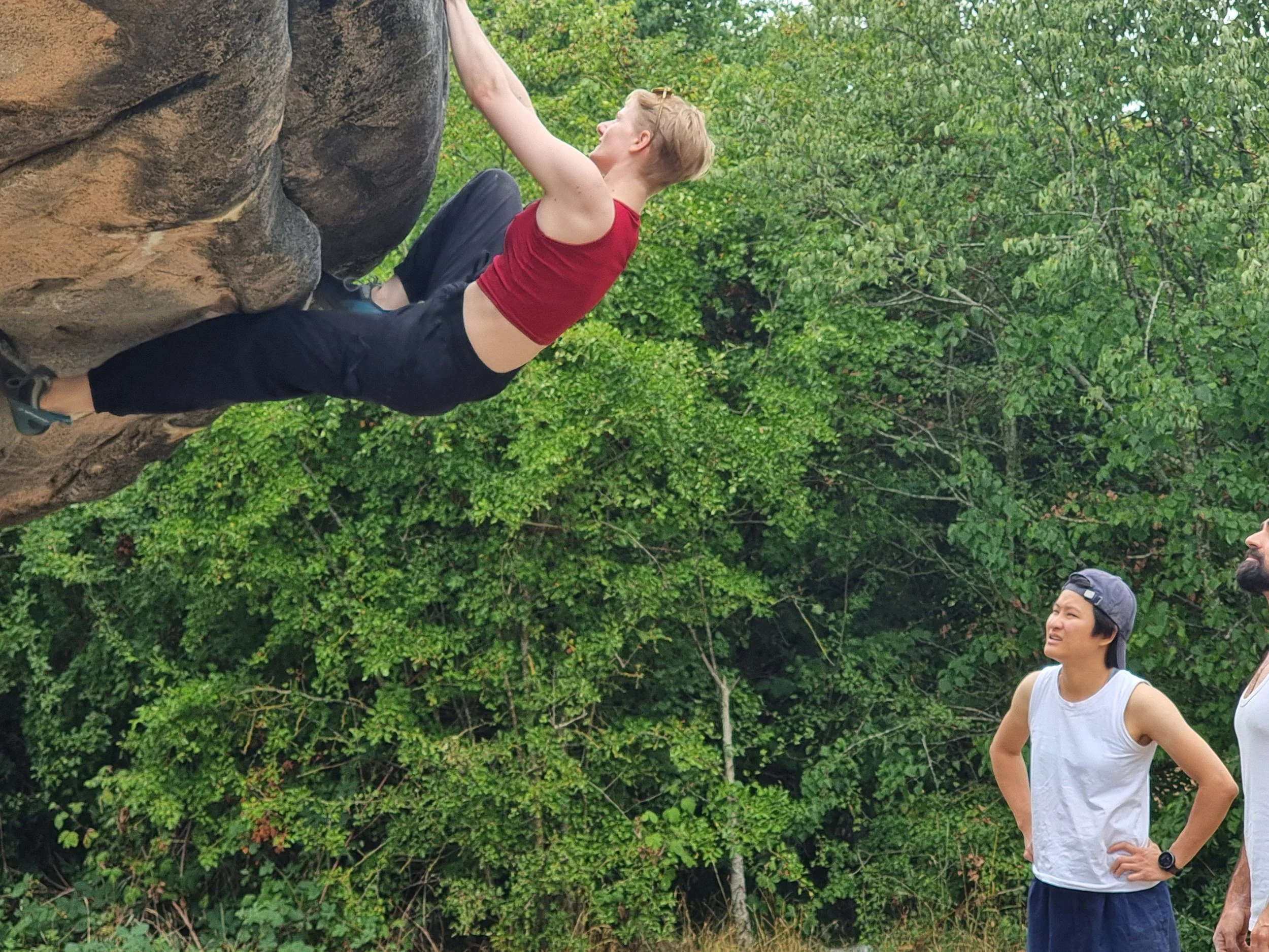 A peron with short blonde hair climbing a rock wall outdoors, wearing a red sleeveless top and black pants, with two people watching them.