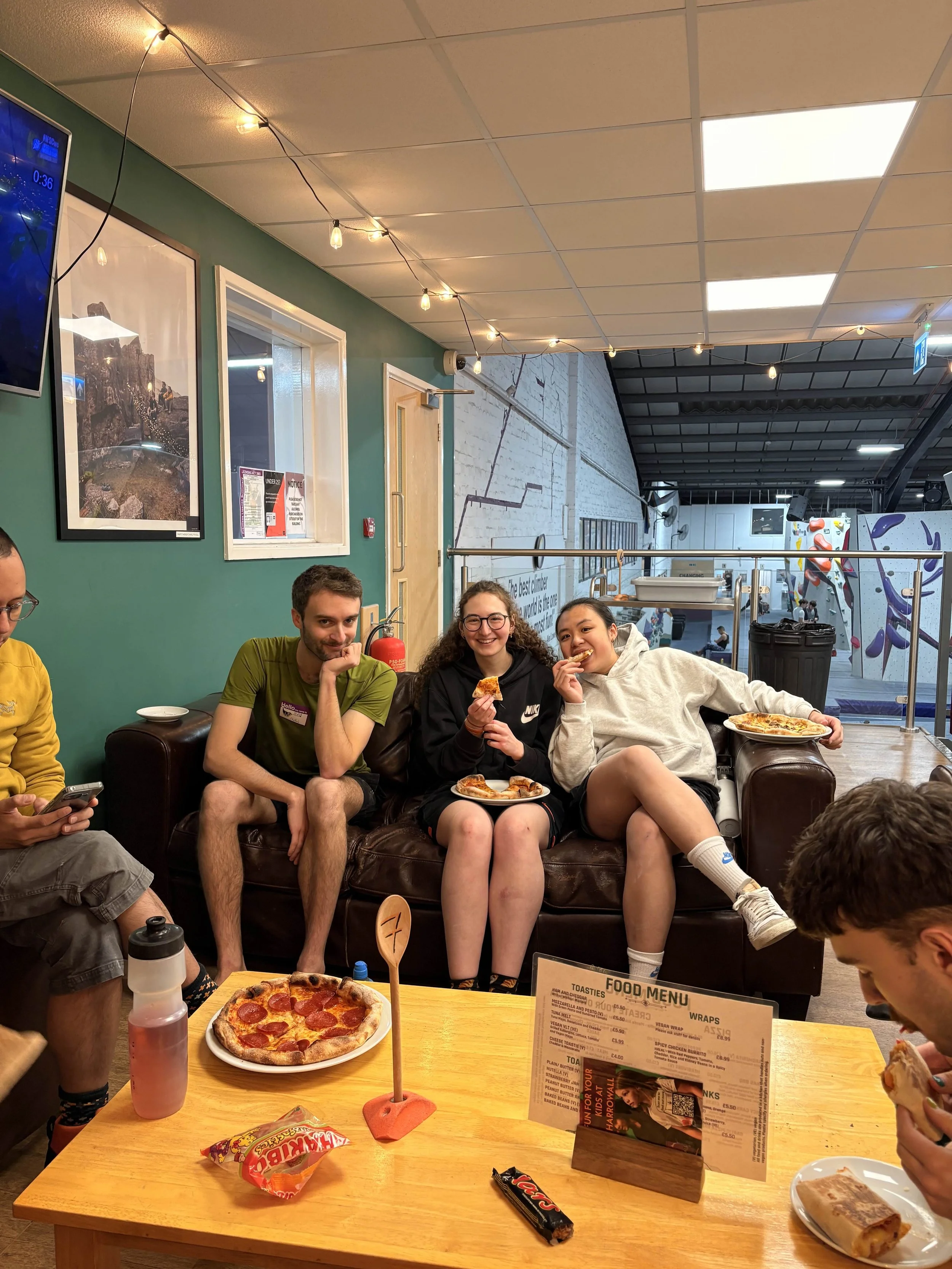 Four young adults sitting on a brown leather couch in a casual restaurant, smiling and enjoying pizza, with a table of snacks and drinks in front of them. The background shows a rock climbing wall, posters, and a food menu on the table.