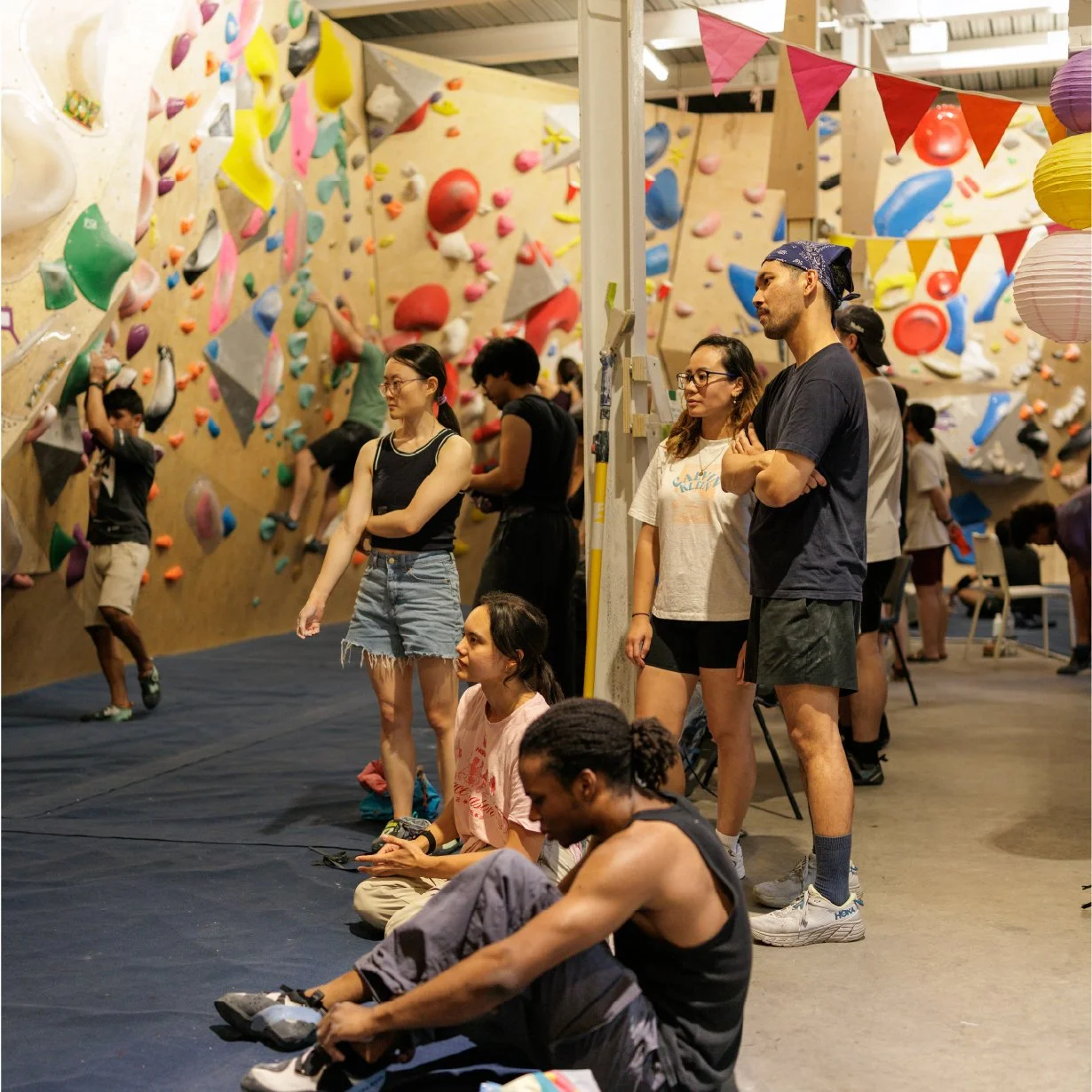 A group of people at an indoor rock climbing gym, watching climbers on a colorful bouldering wall with various holds. Some sit on the floor, others stand, observing and waiting for their turn.