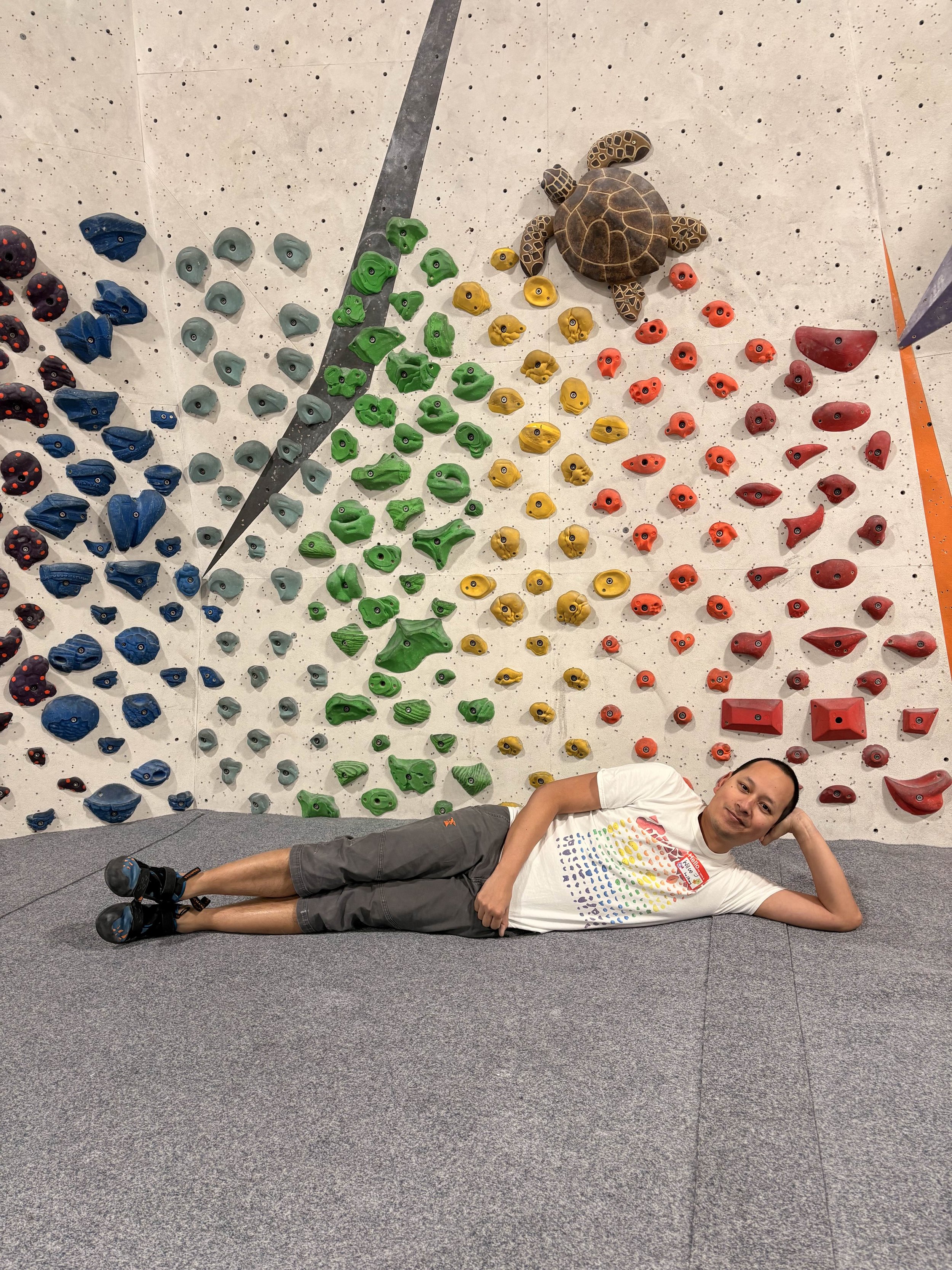 A person lying on his side on a gray carpeted floor in front of a colorful climbing wall. The person is wearing a white t-shirt, gray shorts, and climbing shoes, and they are smiling at the camera.