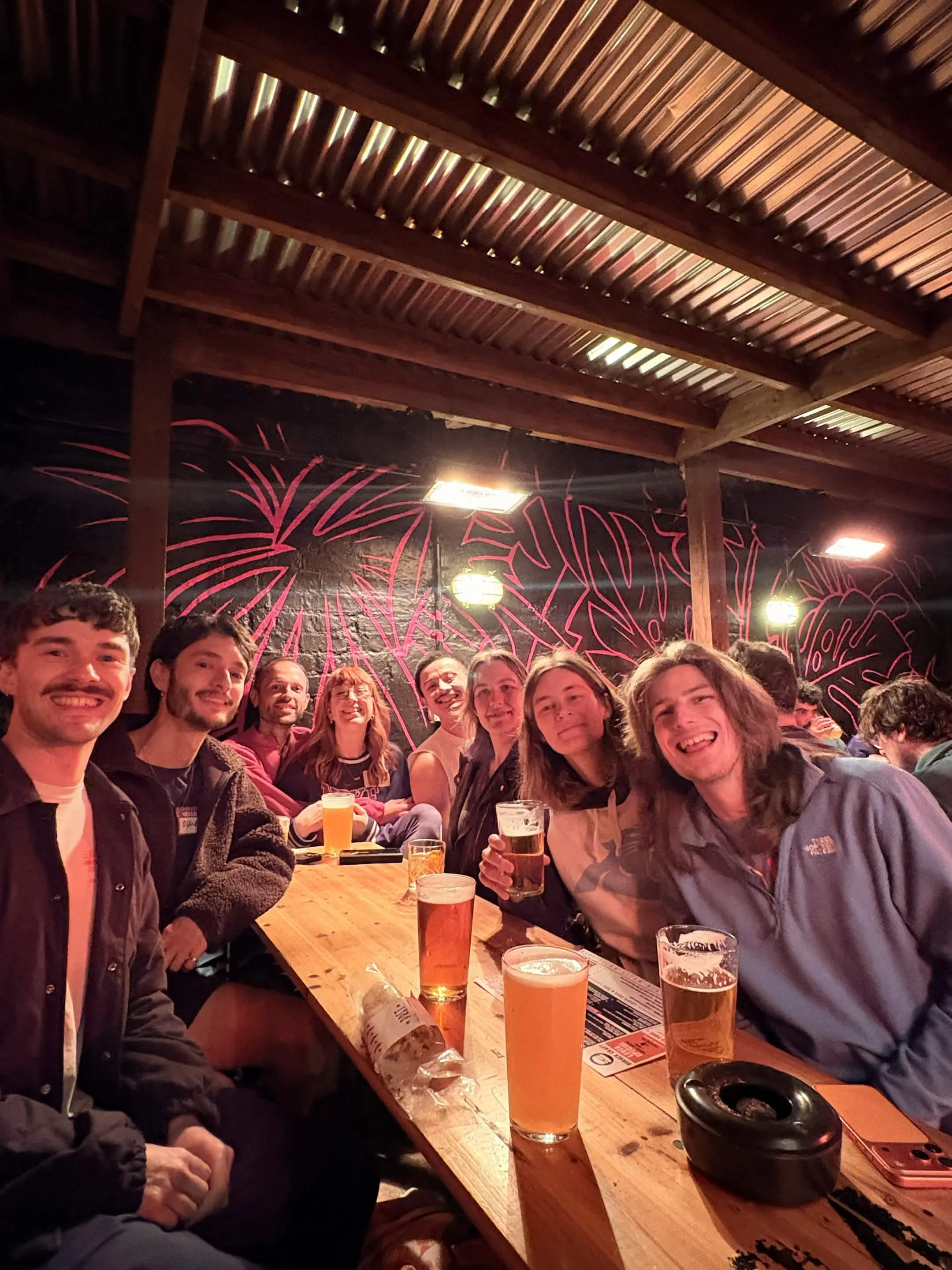 A group of people sitting around a wooden table at a bar or brewery, enjoying drinks and smiling. There are multiple glasses of beer on the table, with some people holding their drinks. The background features a dark wall with red and white firework-like artwork and hanging lights. The ceiling is made of wooden beams and metal roofing.