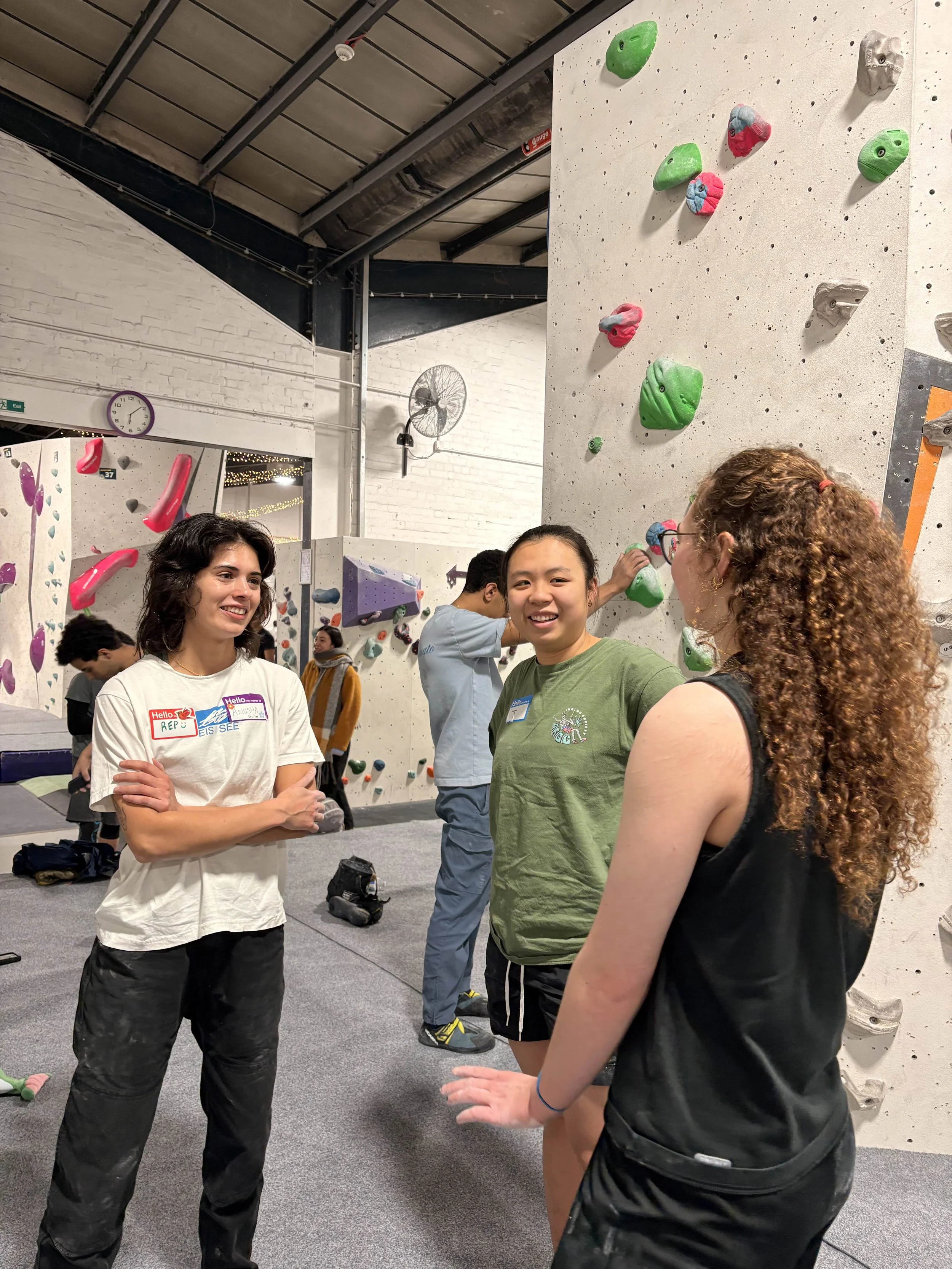 Three young people standing and talking in an indoor climbing gym, with climbing holds on a wall behind them, other people visible in the background.
