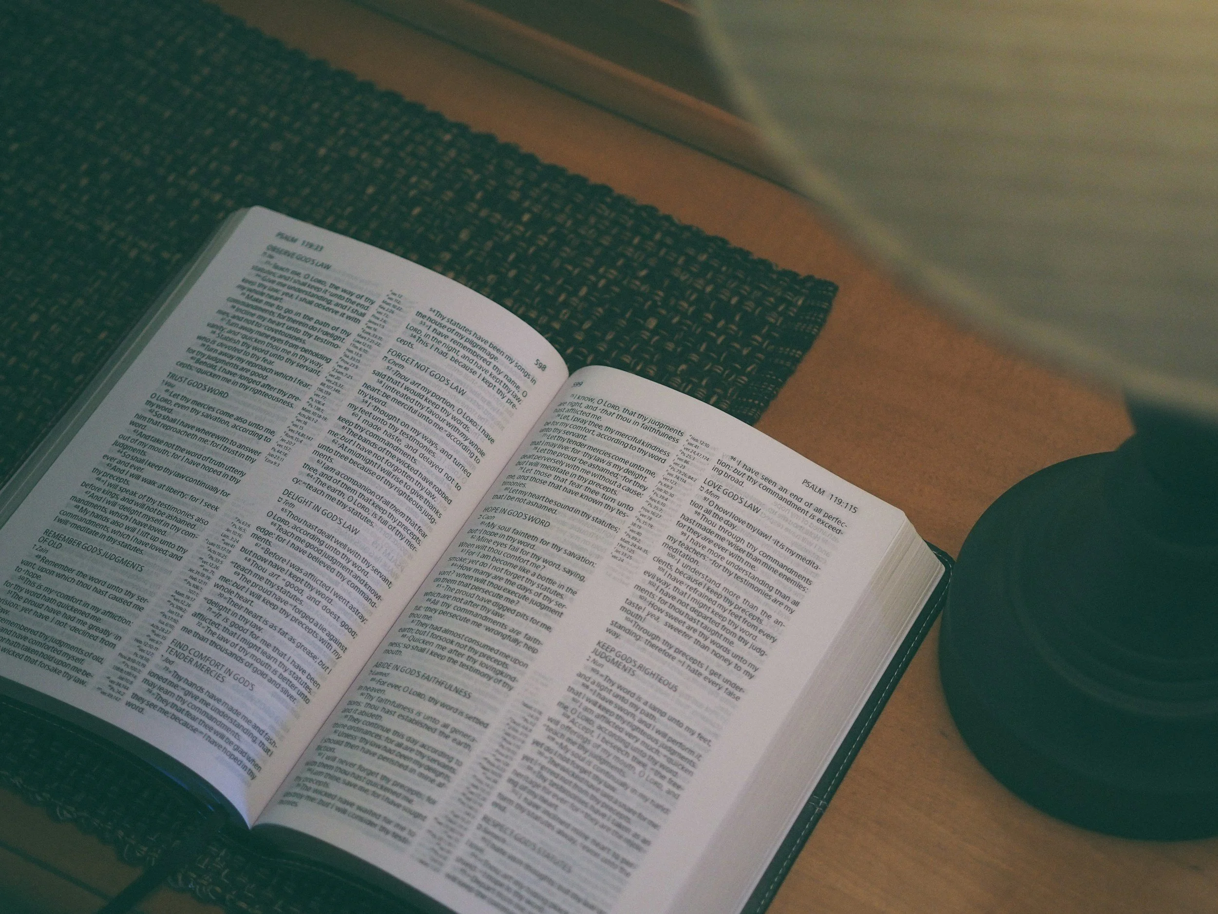 Open Bible on a wooden desk with a dark placemat, and a lamp on the right side.