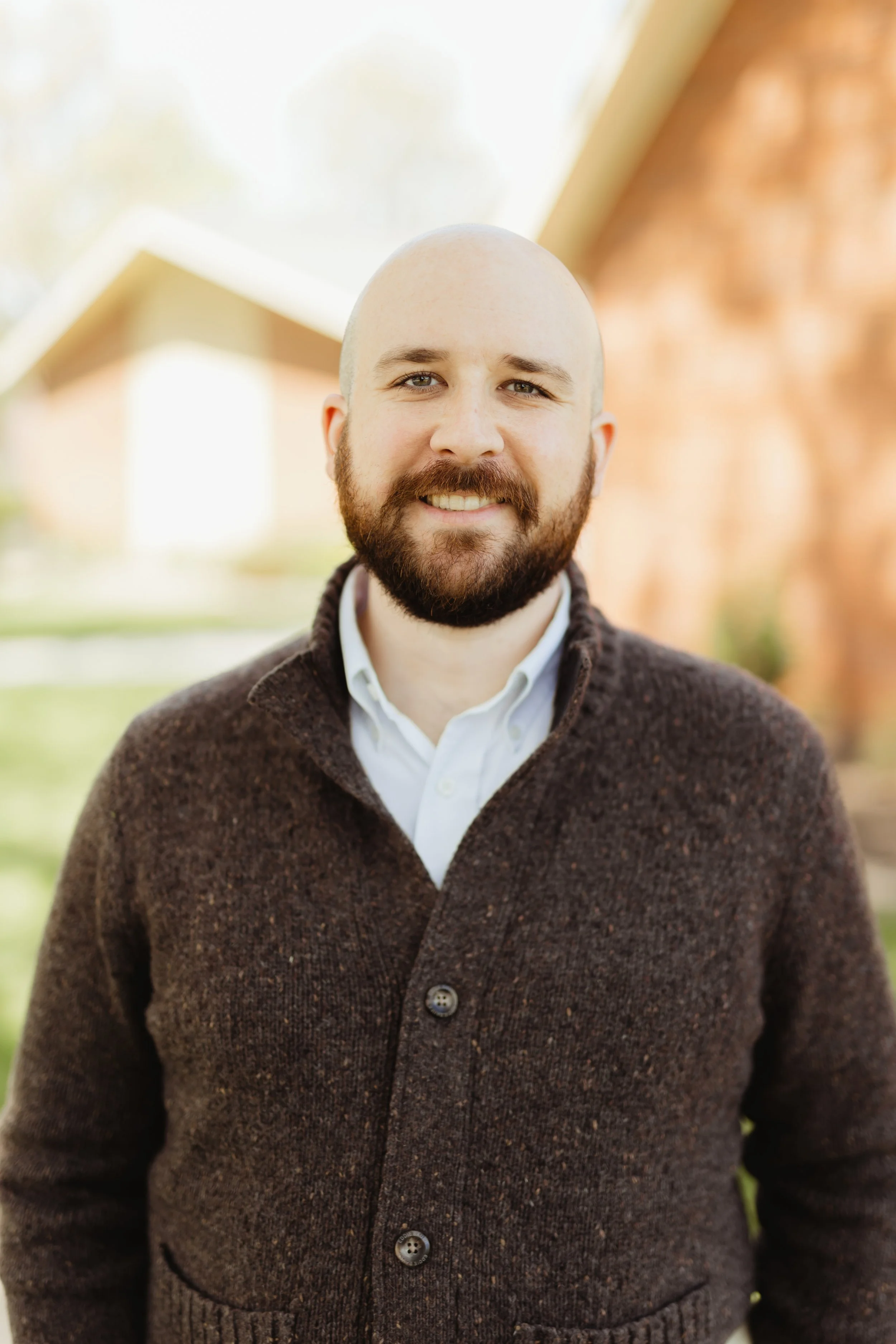 A smiling man with a beard and bald head standing outside in front of a house with a brick wall and gabled roof.