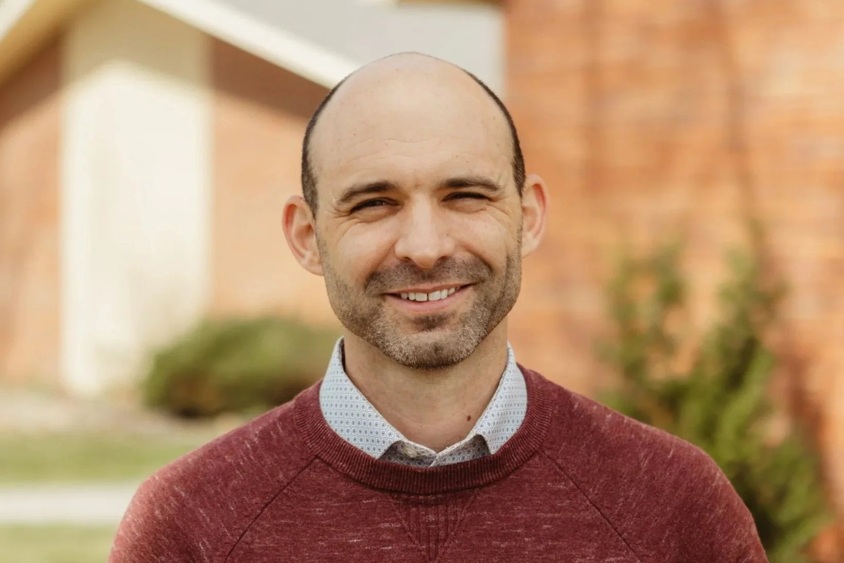 Portrait of a smiling man with a beard and bald head, wearing a white shirt and dark sweater, outdoors with blurred background.