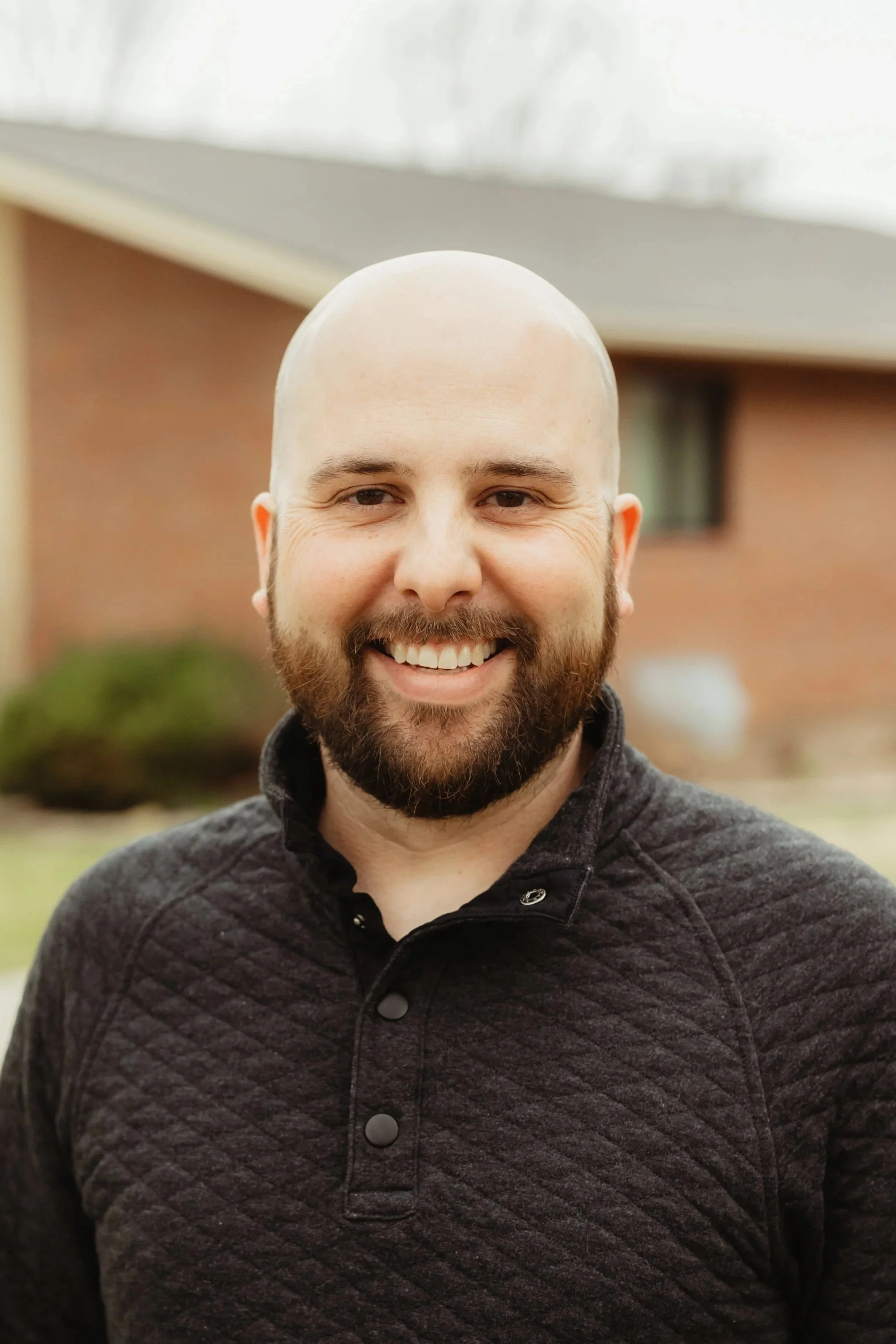 A middle-aged man wearing a navy suit and white shirt, smiling in front of a blurred outdoor background.