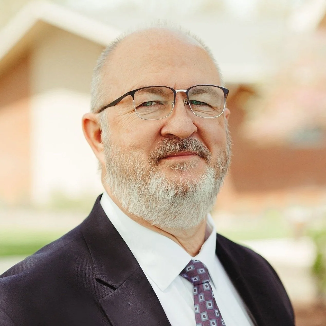 Portrait of a smiling middle-aged man with gray hair and beard, wearing a dark blazer and light blue shirt, outdoors with blurred greenery in the background.