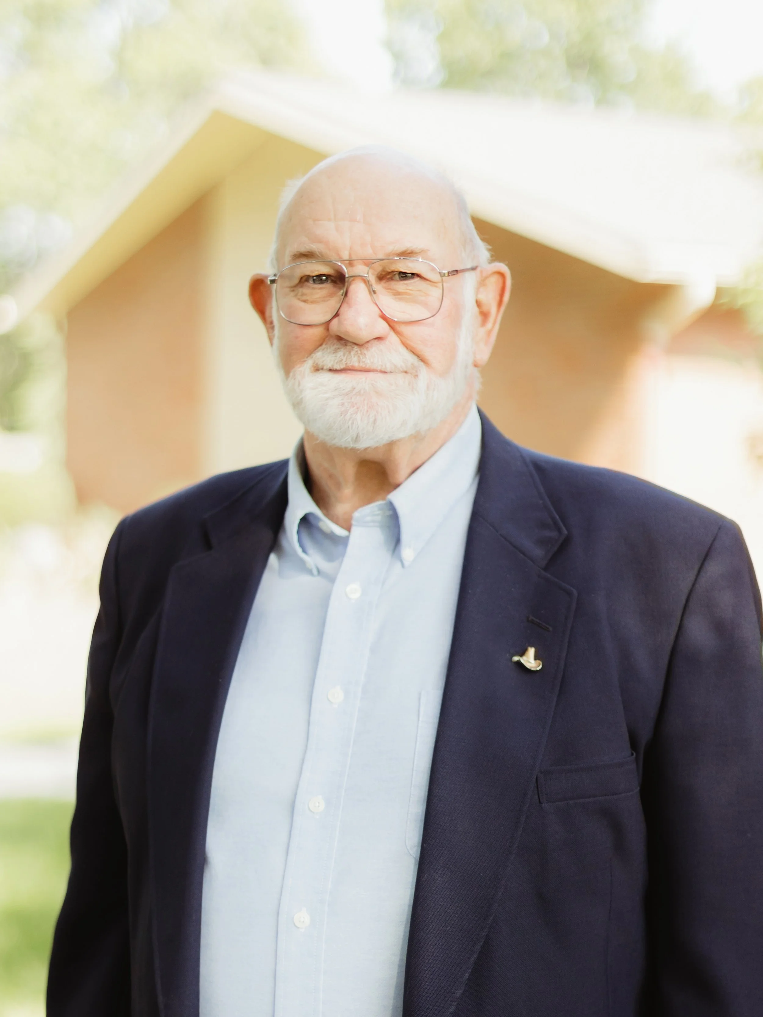 Older man with white hair and beard wearing glasses, a dark blazer, and a light blue shirt, standing outdoors with a house in the background.