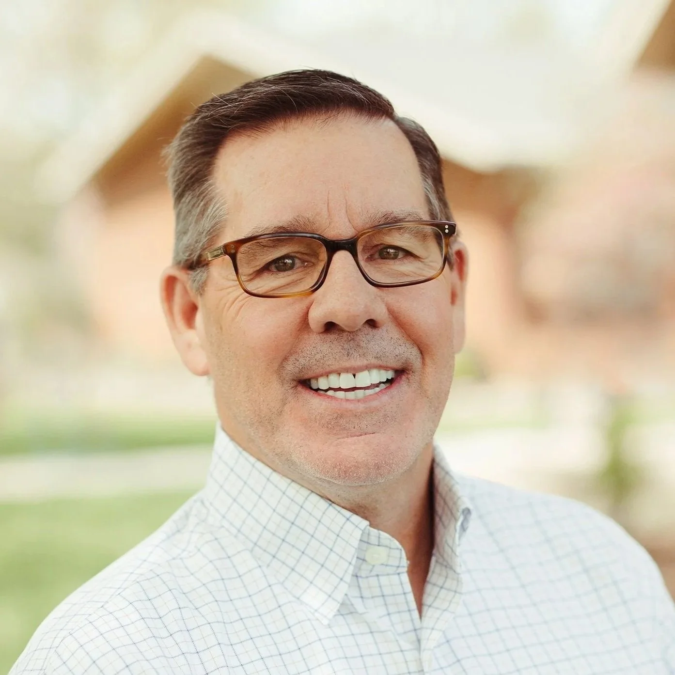 Headshot of a smiling man with a shaved head and beard, wearing a plaid shirt, outdoors in natural light.