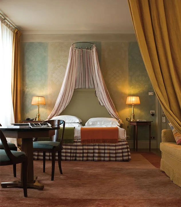 A hotel room with a bed that has a pink canopy and curtains draping down from the ceiling, two bedside lamps, a small table, a wooden desk with a phone, and a cushioned armchair.