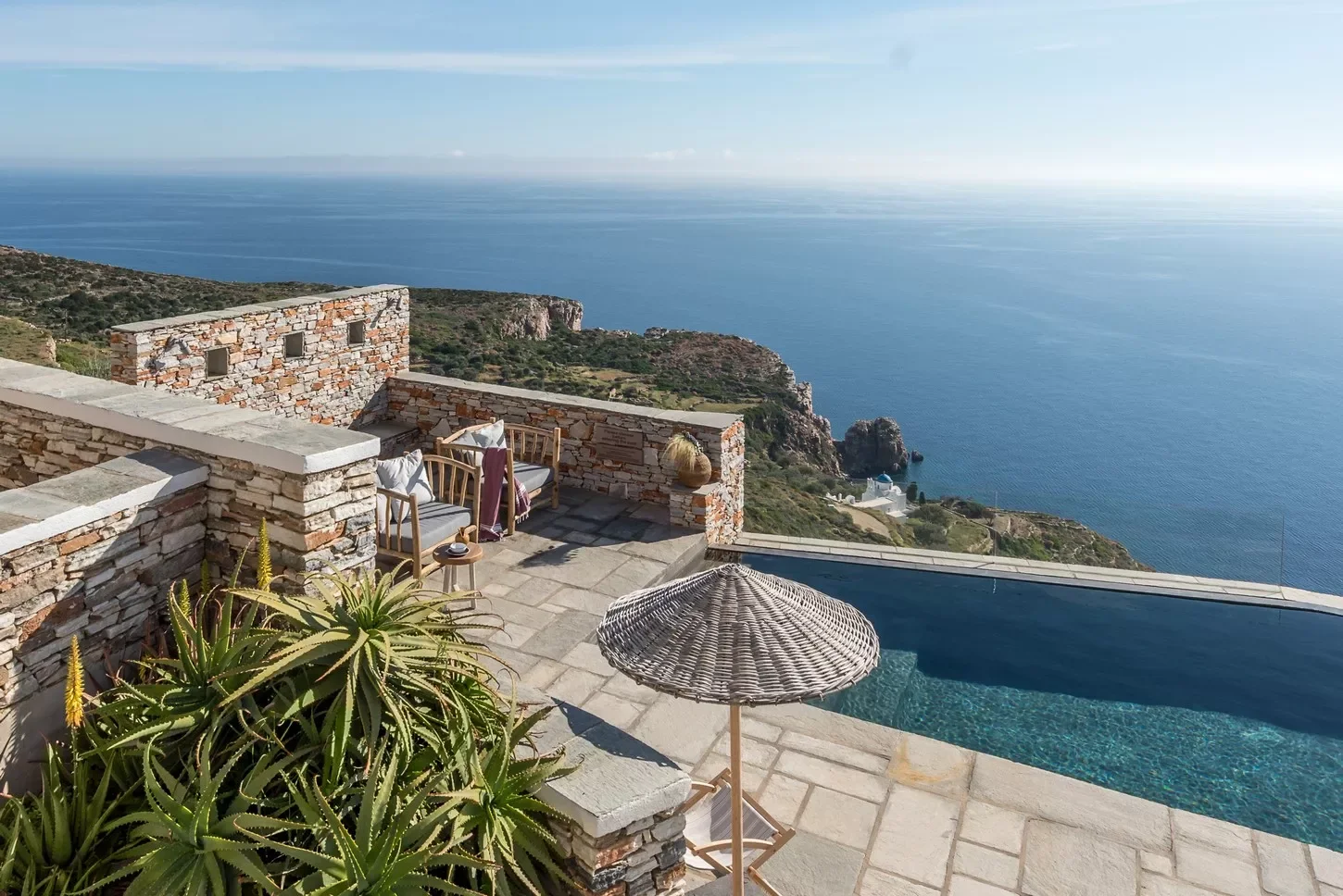 View from a terrace in Greece with a small pool overlooking the ocean, with stone walls, outdoor furniture, and a straw umbrella, overlooking cliffs and the sea.