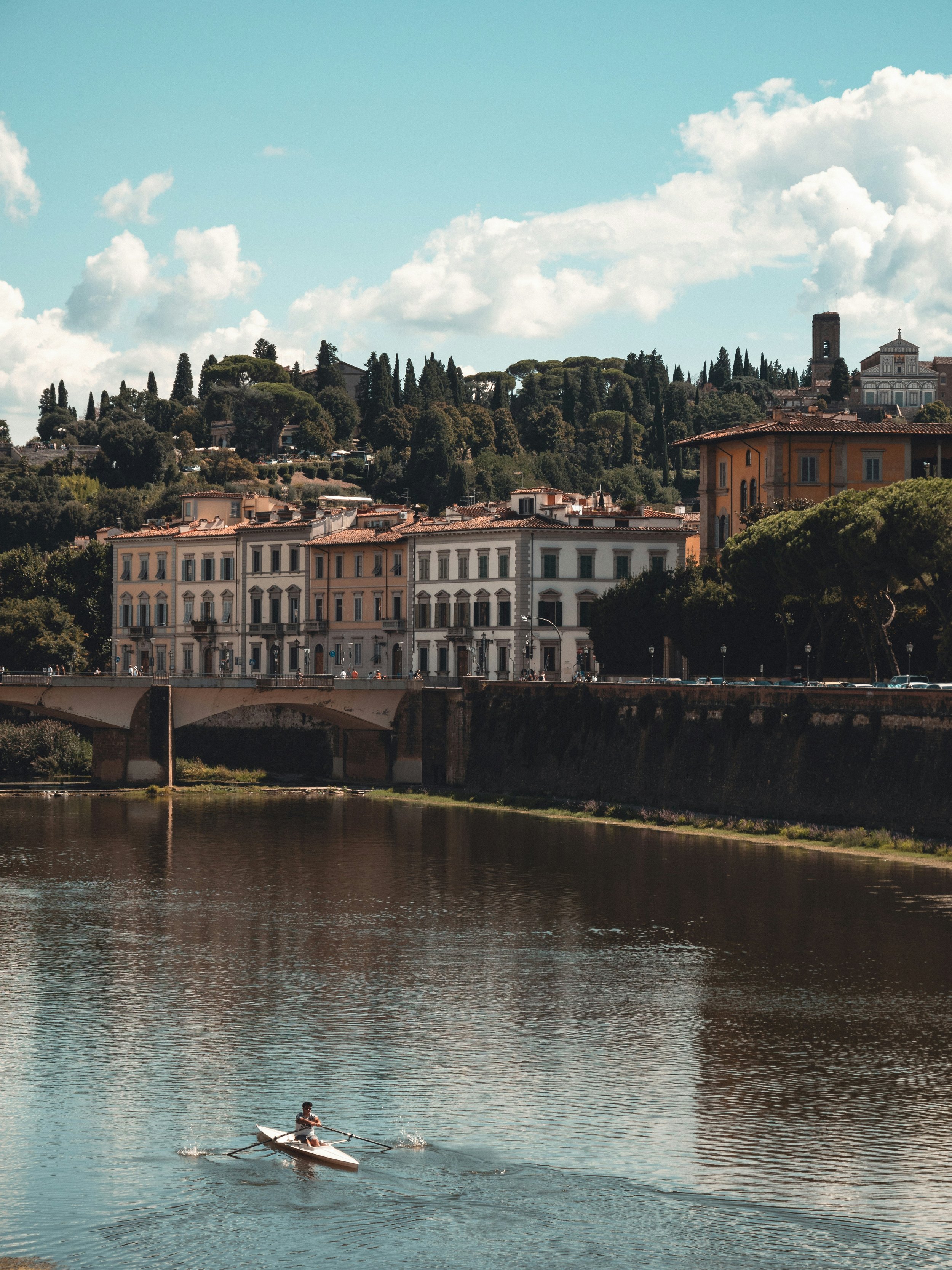 A person rowing a boat on a river in front of a hillside with colorful historic buildings and lush green trees, under a partly cloudy sky.
