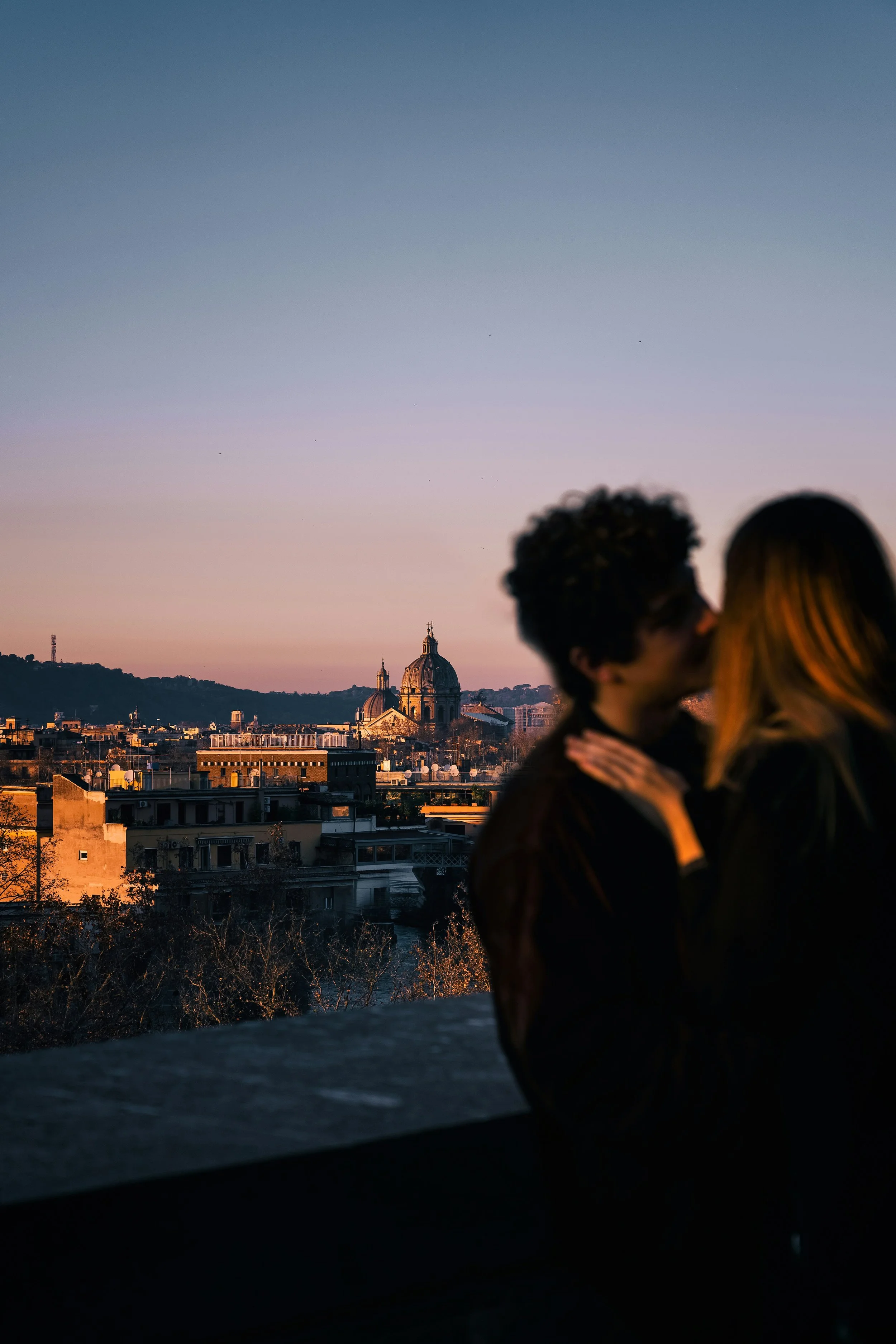 A couple kissing on a rooftop with a city skyline in Rome with St. Peter's Basilica in the background during sunset.