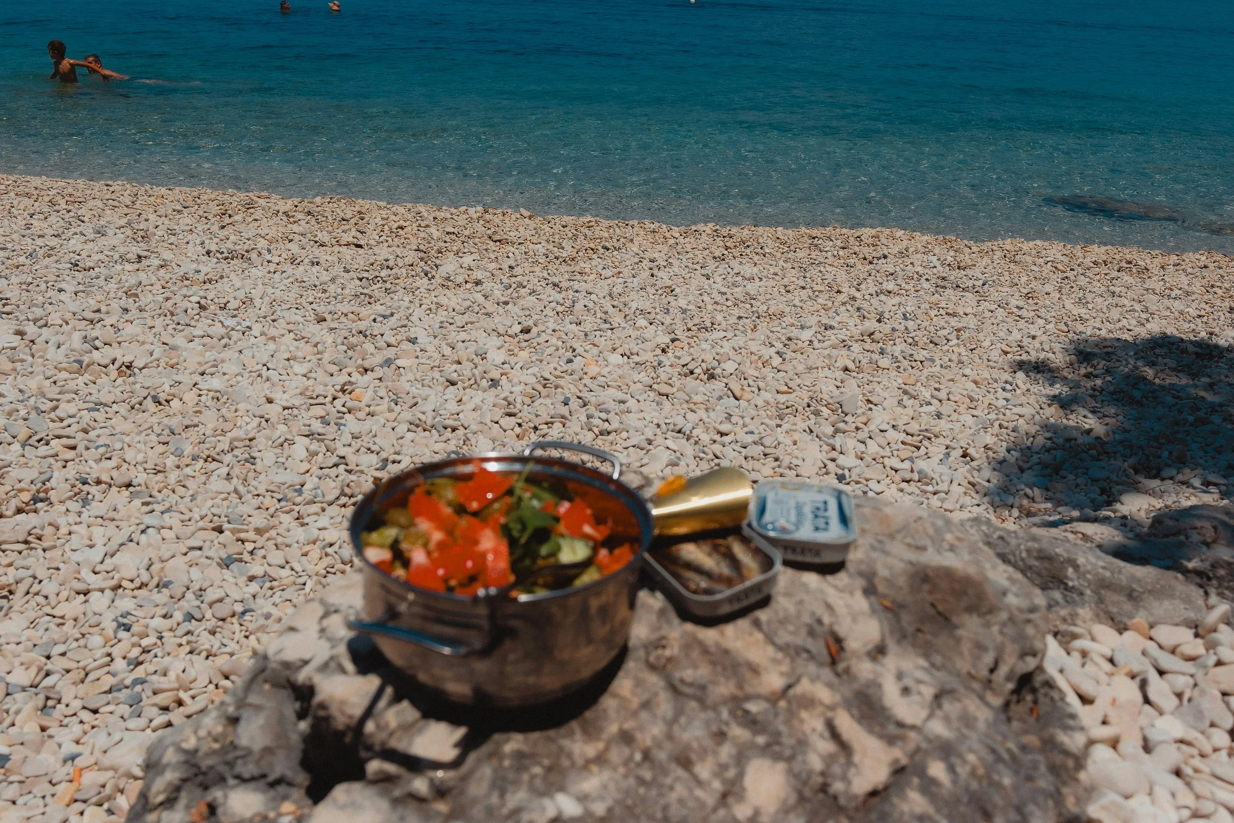 A stove with a pot of salad, canned food, and grill on a large rock at a pebble beach, with the ocean and swimmers in the background.