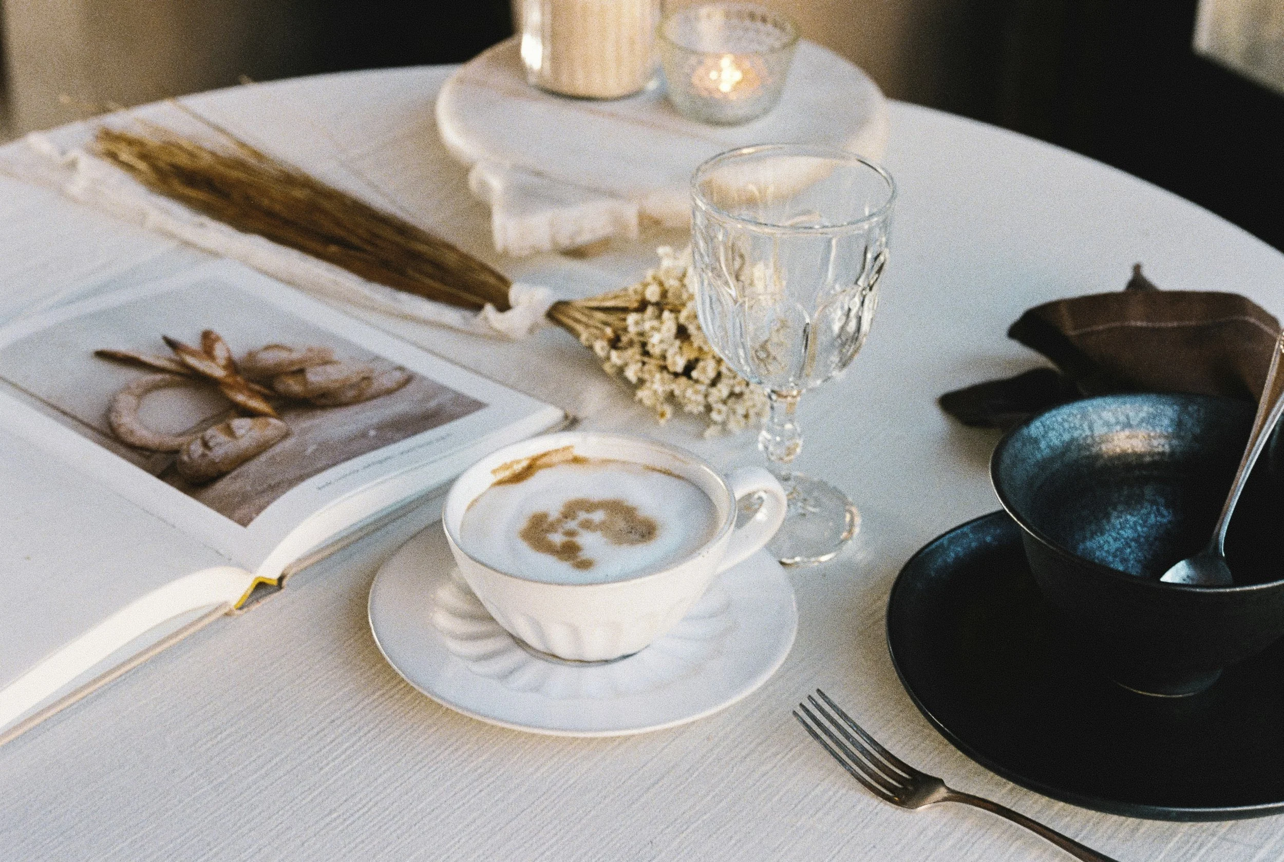 A table setting with a cup of coffee, an empty glass, a black bowl with a spoon, and an open book with a photograph of shrimp, on a white tablecloth.