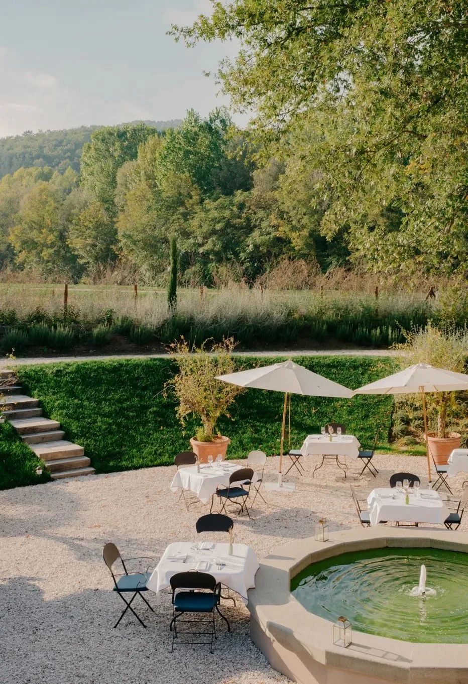 Outdoor dining area with tables covered in white tablecloths, black chairs, large white umbrellas, potted plants, and a fountain, set against lush green trees and distant hills.
