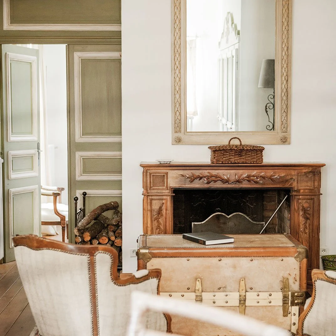 A cozy living room with a vintage wooden fireplace mantel, a large mirror above it, a wicker basket on the mantel, a chest in front, a fireplace tool, a beige armchair, and a stash of firewood.