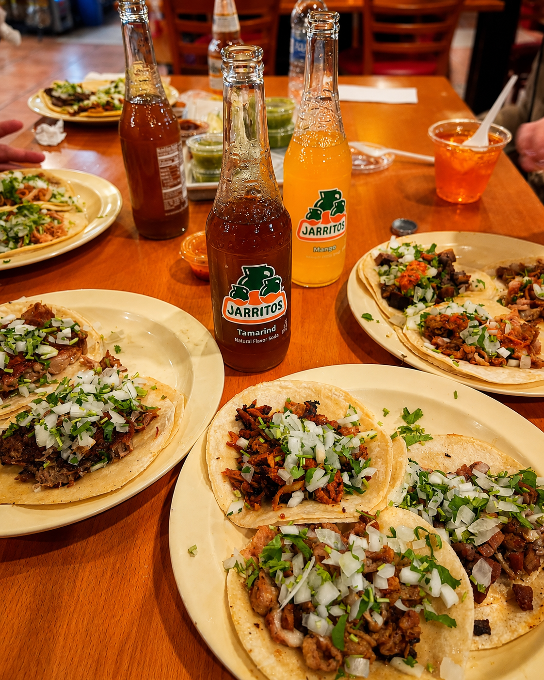 A table with several plates of tacos topped with chopped onions, cilantro, and meat, along with bottles of Jarritos soda in tamarind and mango flavors, in a restaurant setting.