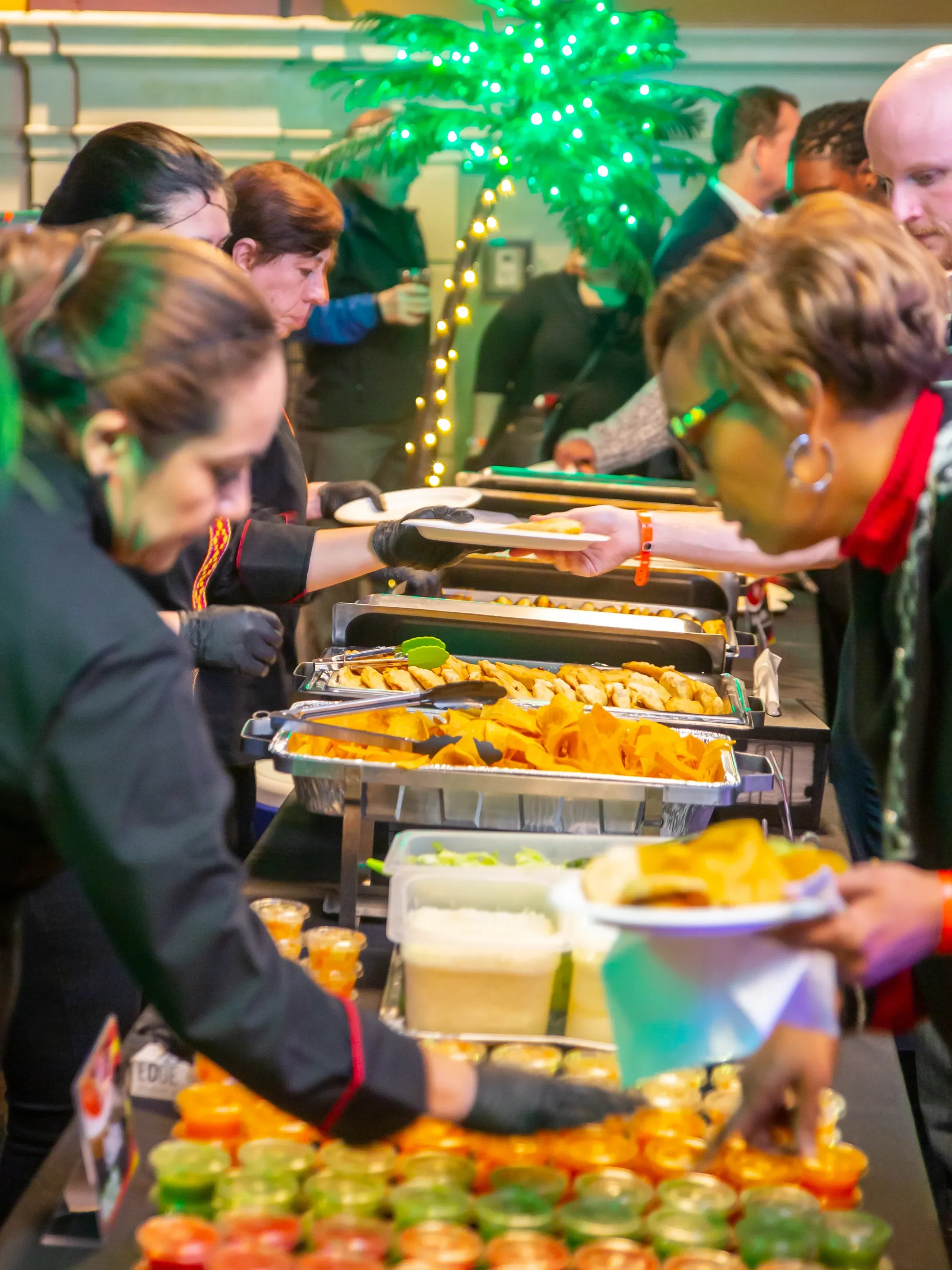 People serving and selecting food at a buffet table with various dishes, including vegetables and appetizers, under green and yellow Christmas lights, with a decorated tree in the background.