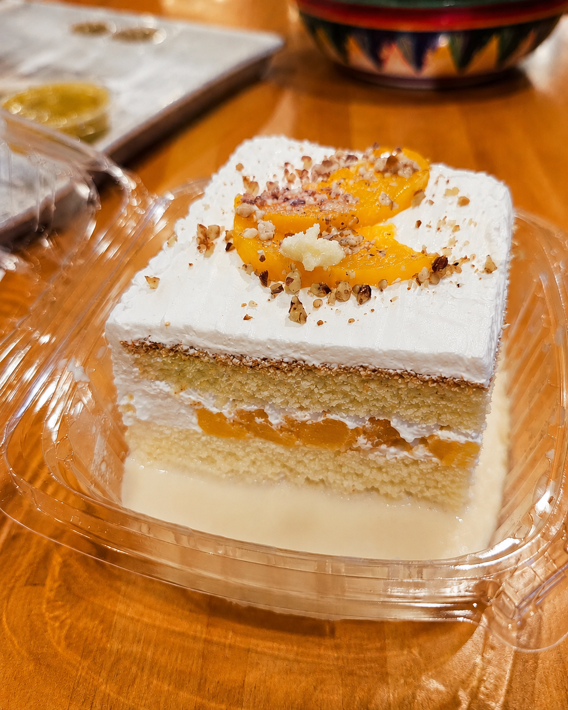 A square slice of layered cake with white frosting, topped with sliced peaches and chopped nuts, in a clear plastic container on a wooden surface.