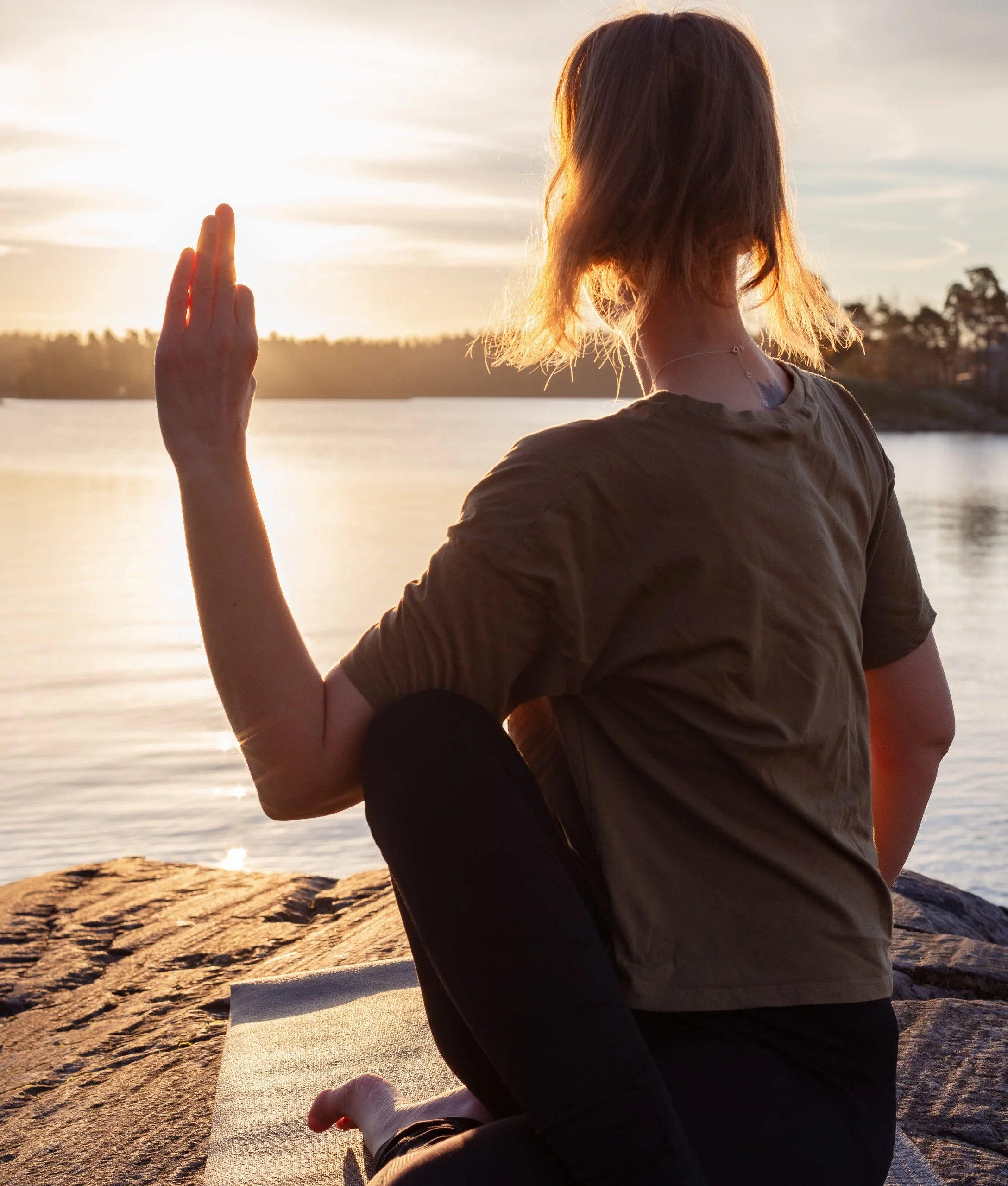 A woman practicing yoga or meditation by the water at sunset, sitting on a mat on rocks with her hand raised in a mudra gesture.
