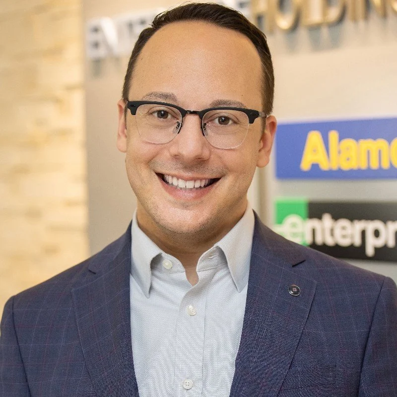 Man wearing glasses and a suit, smiling in front of a wall with Enterprise and Alamo logos.  Career path and leadership journey for resilient individuals wanting to grow.