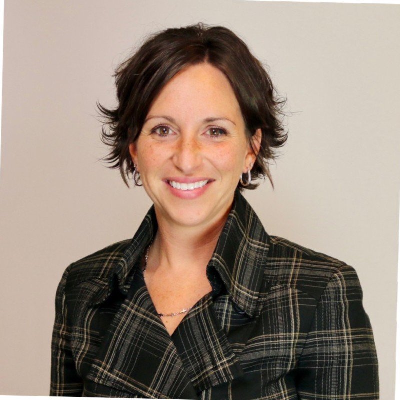 Woman smiling, wearing a plaid jacket with short hair, against a neutral background.  Professional development and leadership coaching drives success.