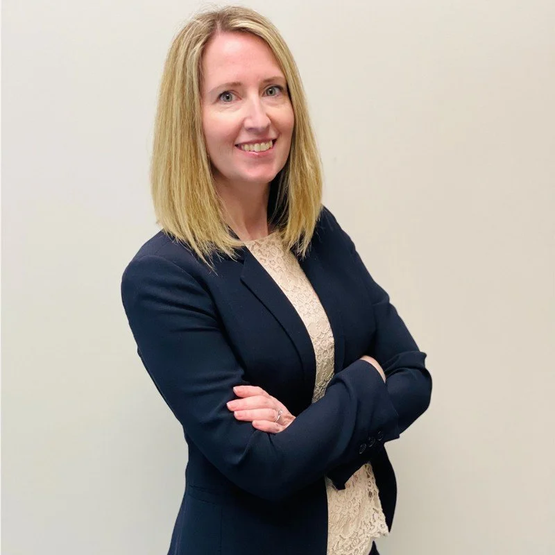 Professional woman in business attire with blond hair, arms crossed, smiling, against a light background.  Female leadership and executive presence.  Confident leader.