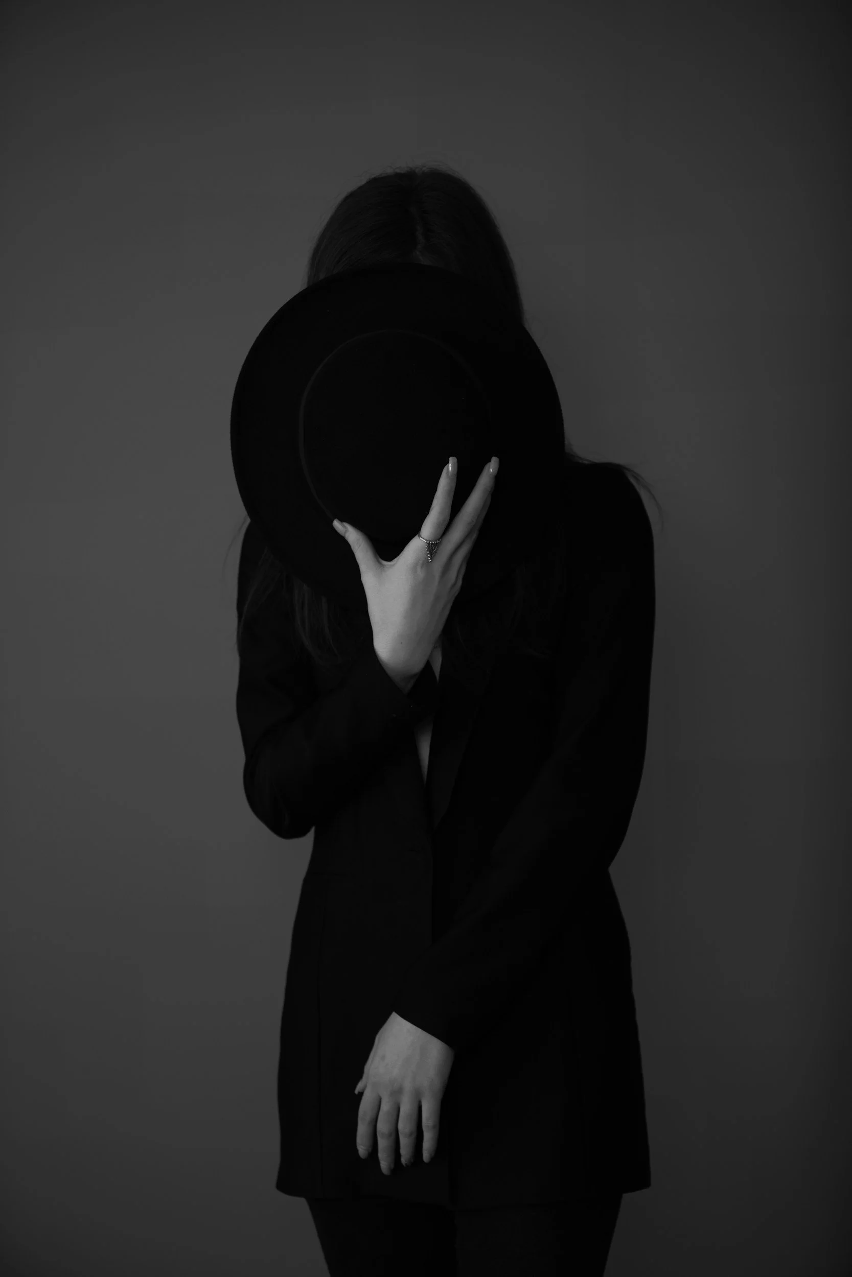 A woman dressed in black, holding a black hat in front of her face, obscuring her identity, in a black and white photo.
