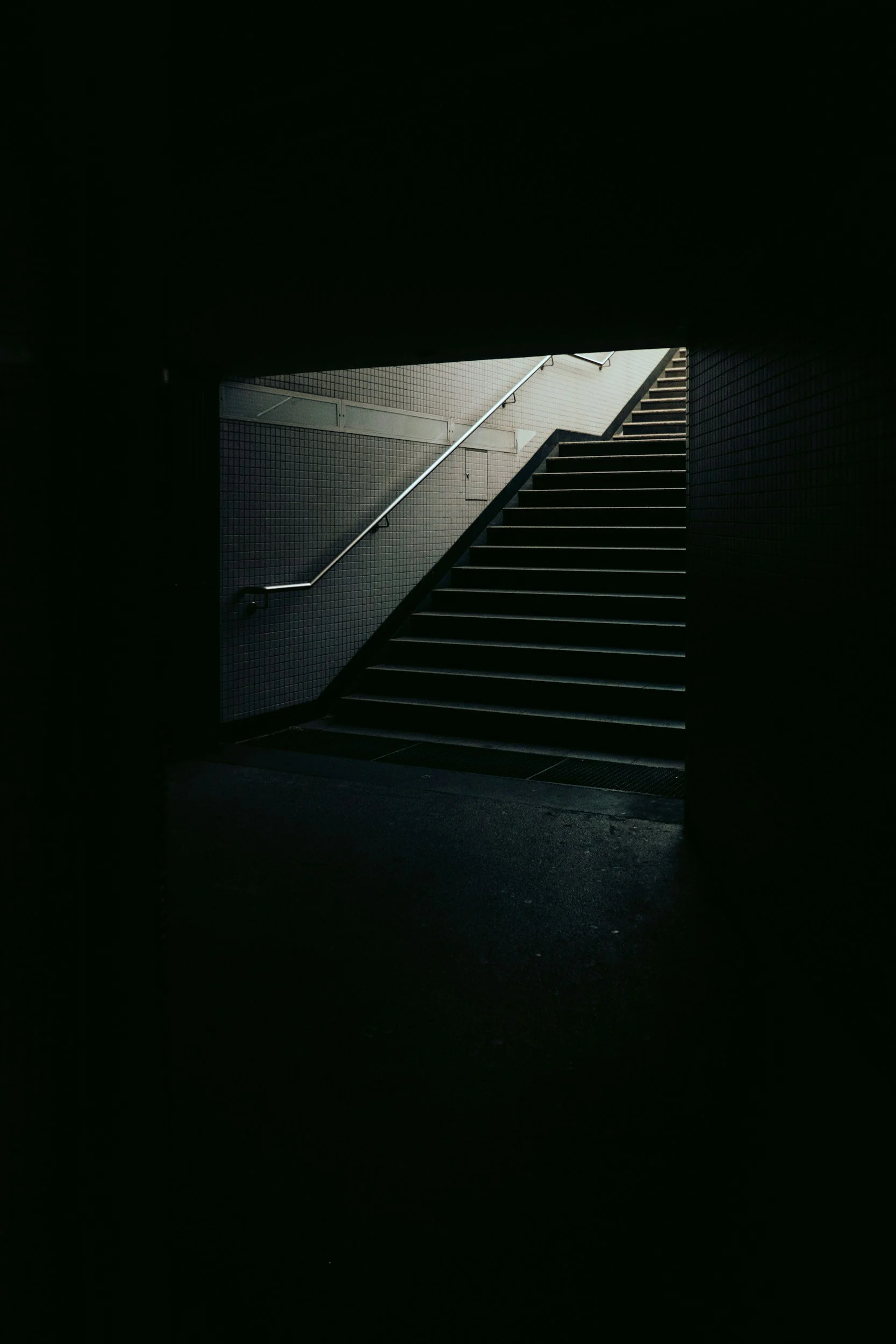 Dark underpass leading to outdoor stairs with metal handrail, illuminated at the top.