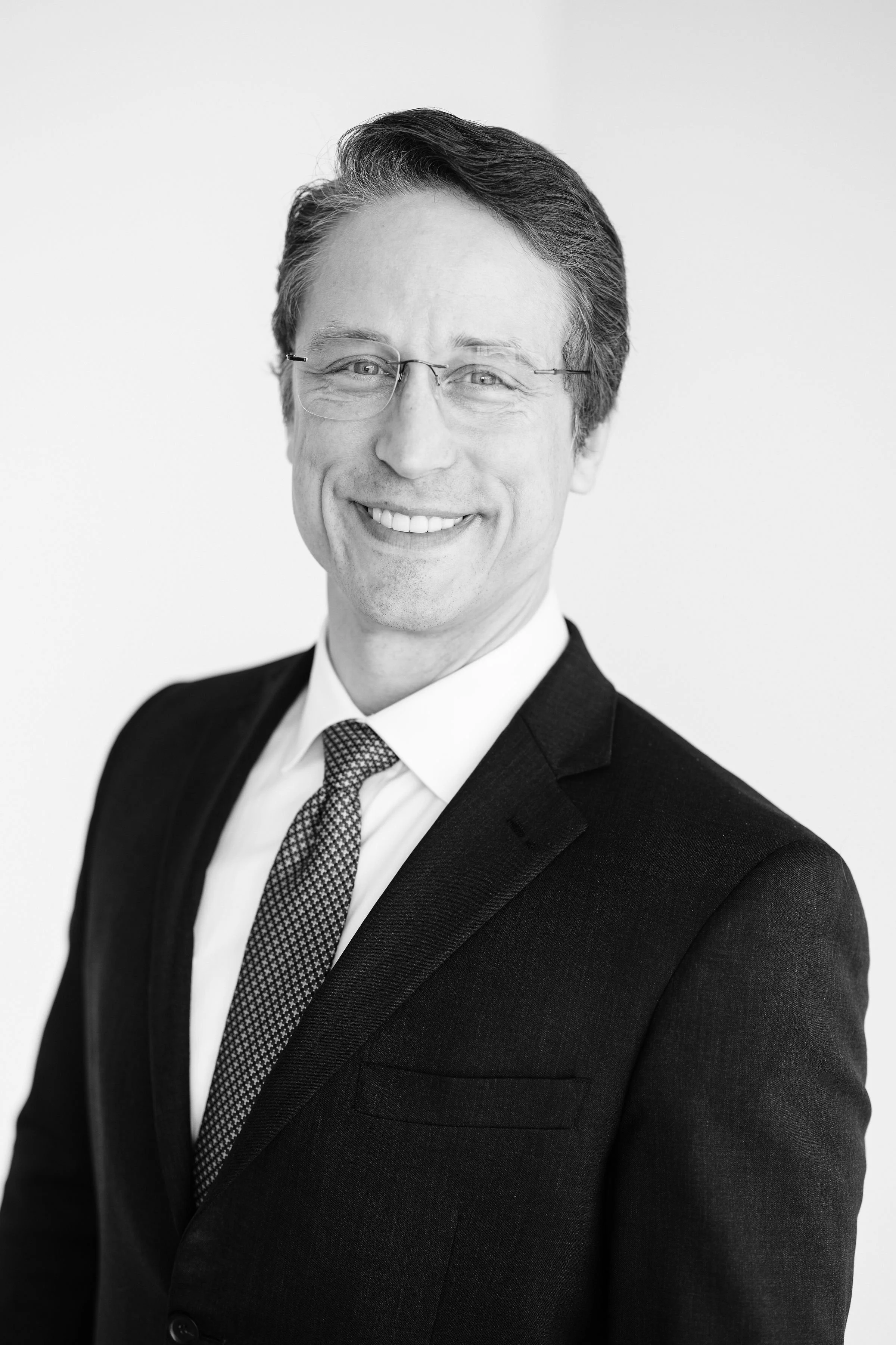A man in a gray suit with a white shirt, purple striped tie, and glasses, standing with his arms crossed against a plain background.