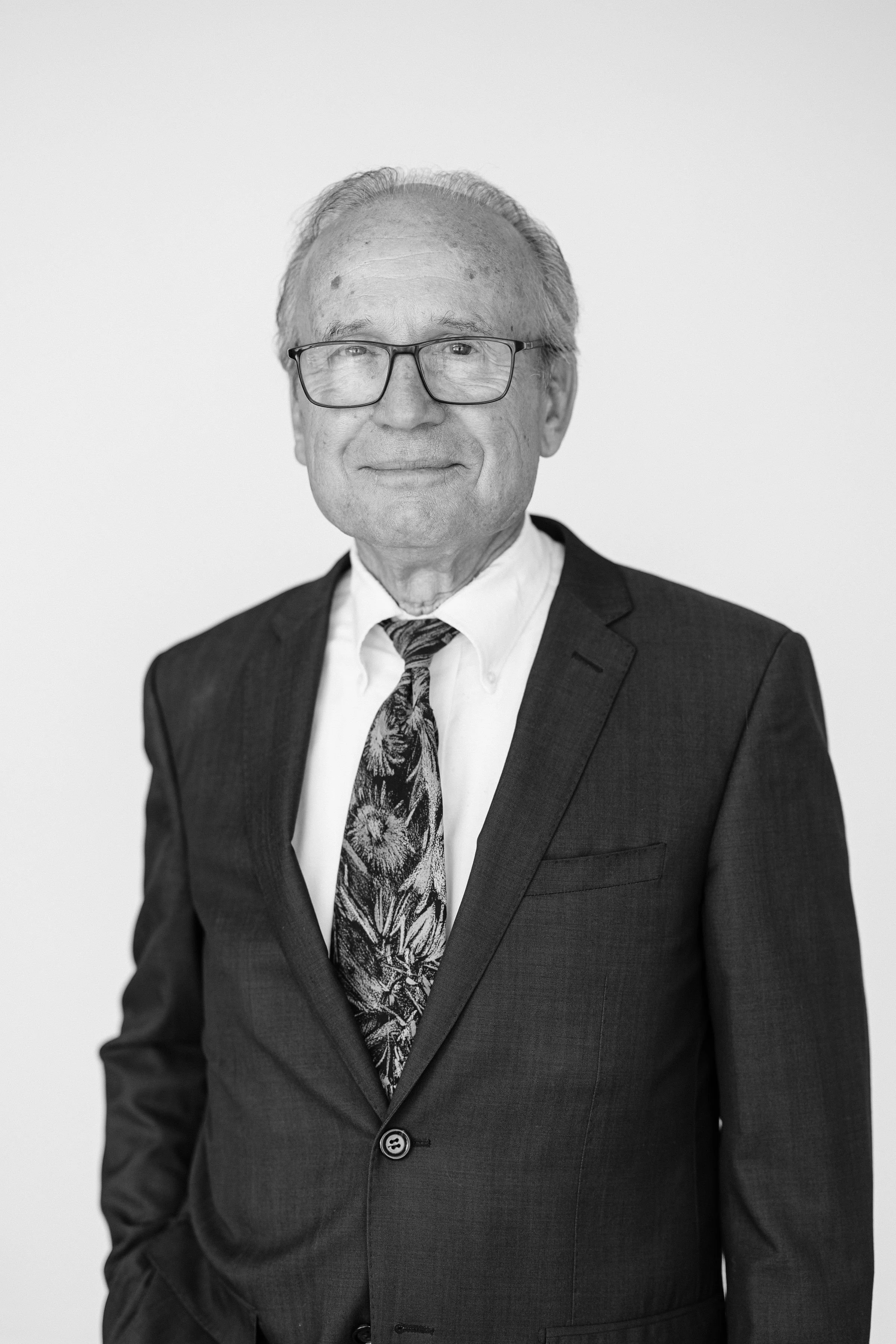 Smiling middle-aged man in glasses wearing a blue suit, white shirt, and blue tie, standing against a plain white background.