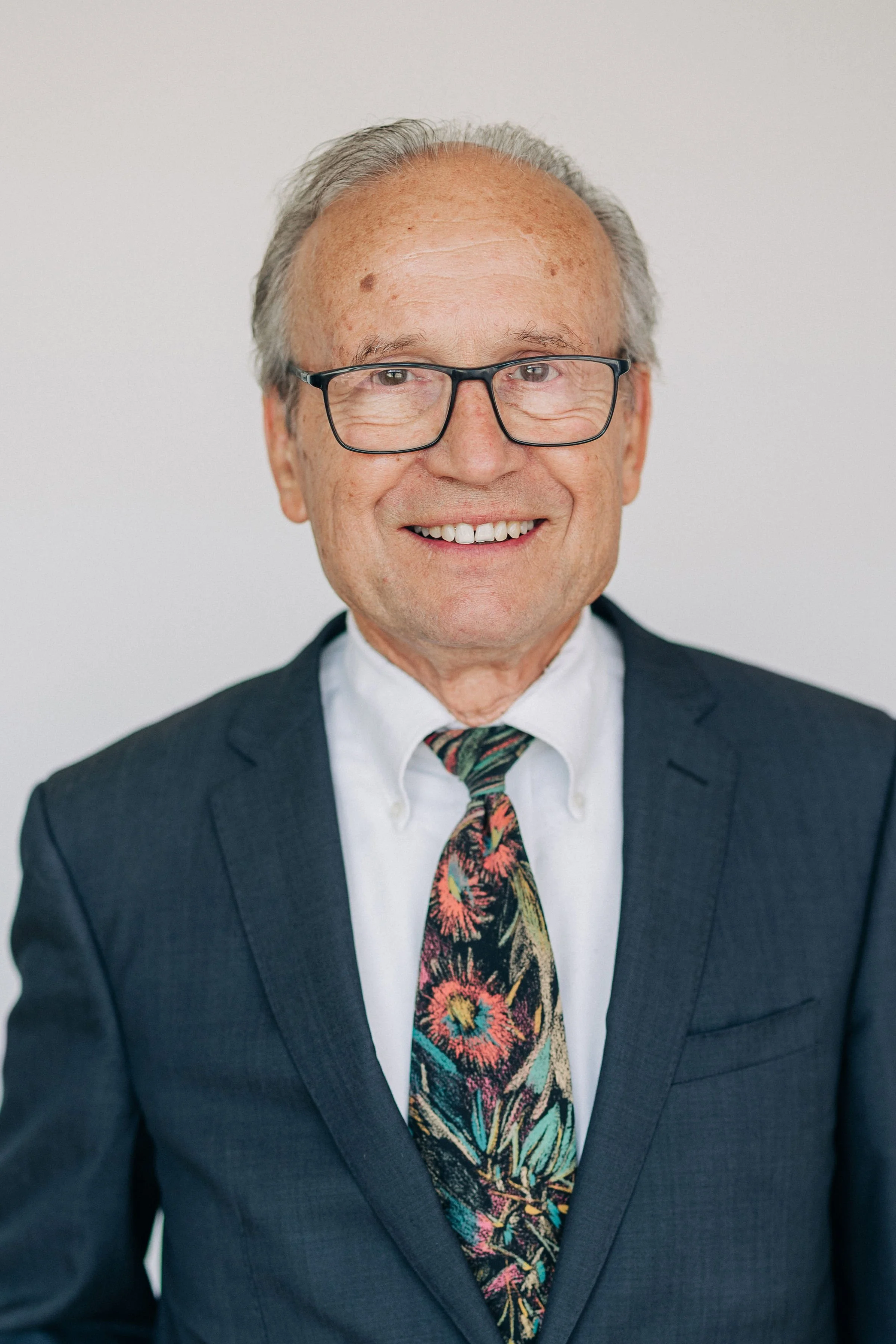 A smiling middle-aged man in a dark suit, white shirt, and blue tie, with glasses and a gold watch, standing with arms crossed against a plain light background.