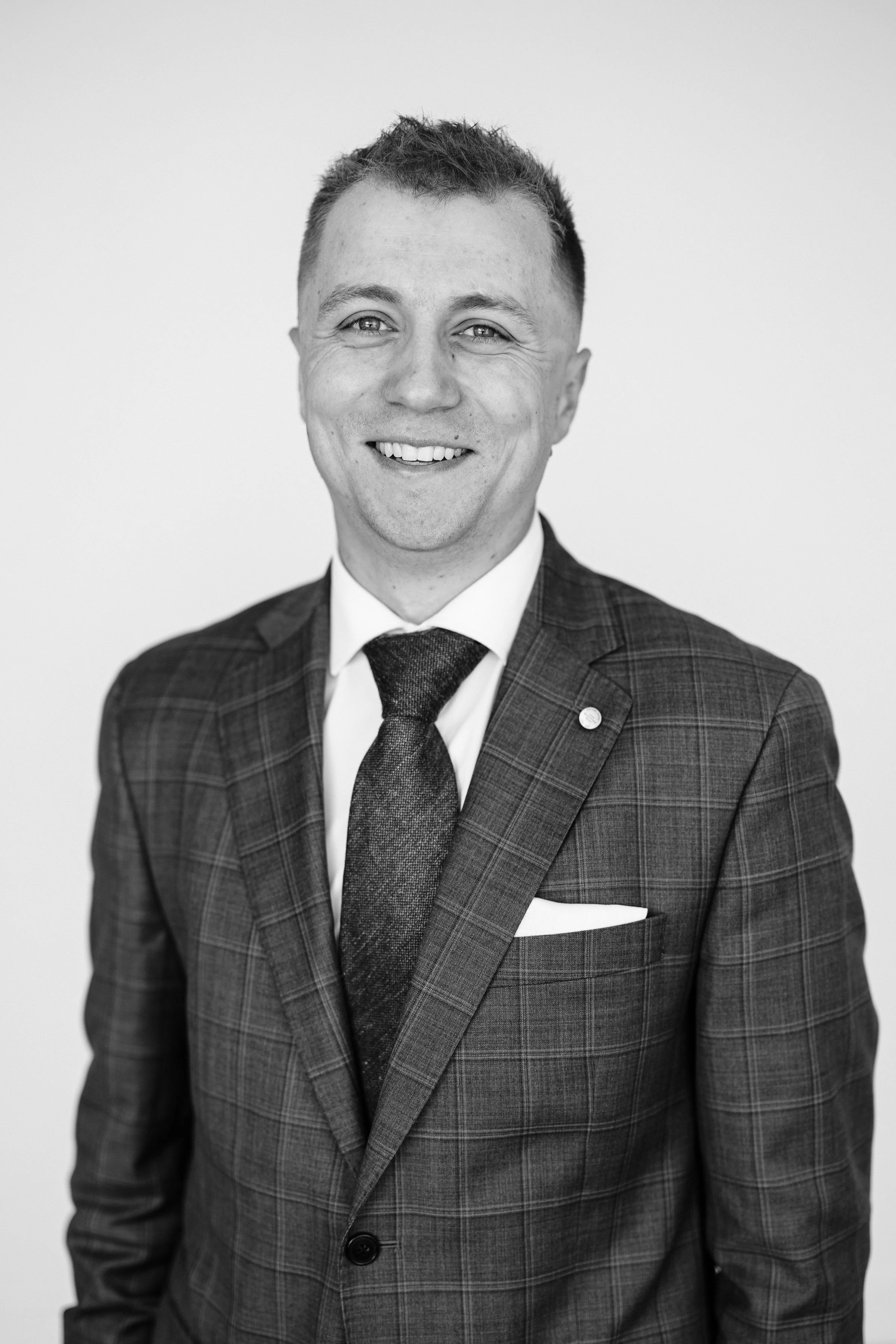 A young man with light brown hair, blue eyes, and fair skin dressed in a black suit, light blue shirt, and black tie, smiling at the camera against a plain white background.