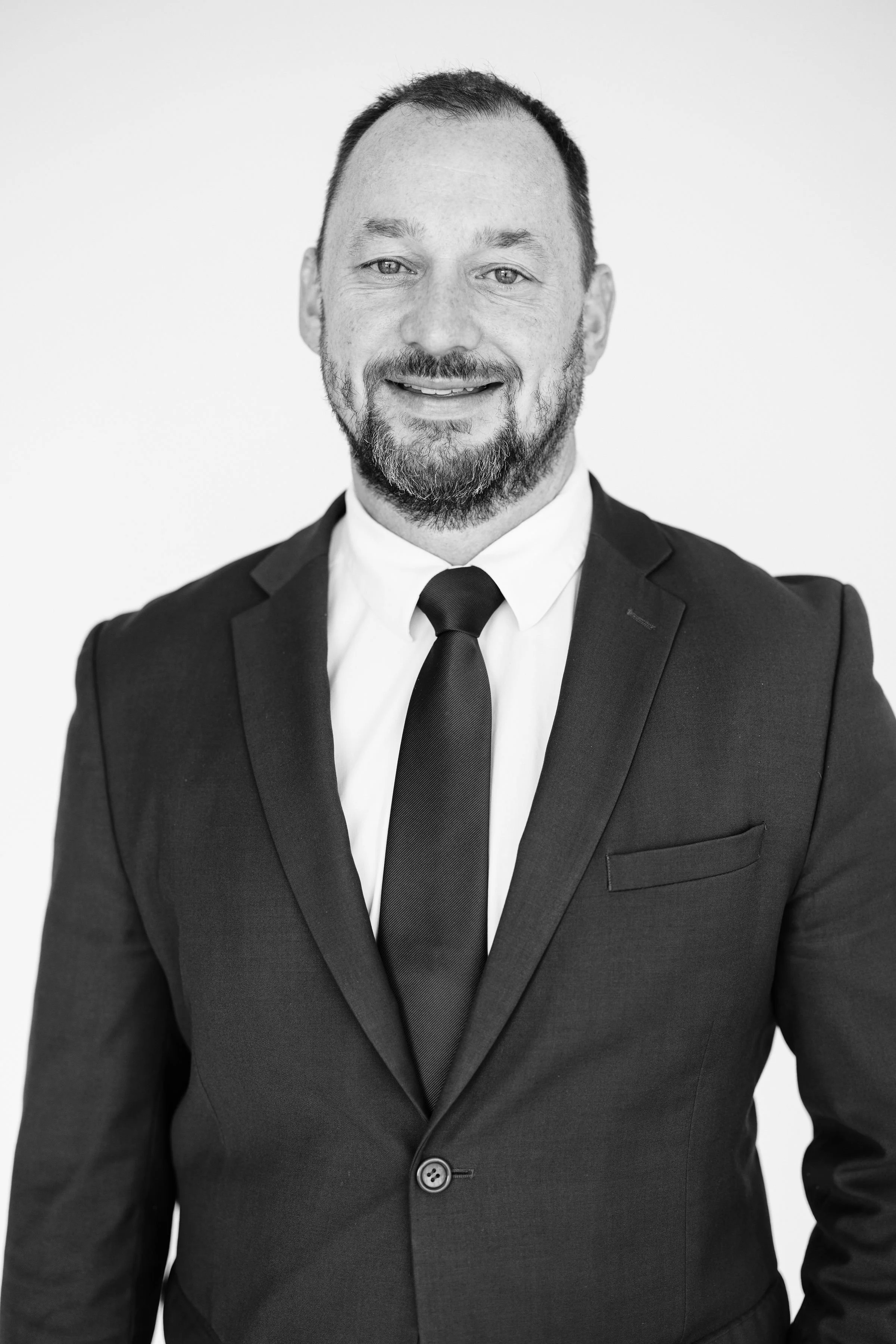 A man in a blue suit sitting in a black office chair, smiling, with a plain white wall background.