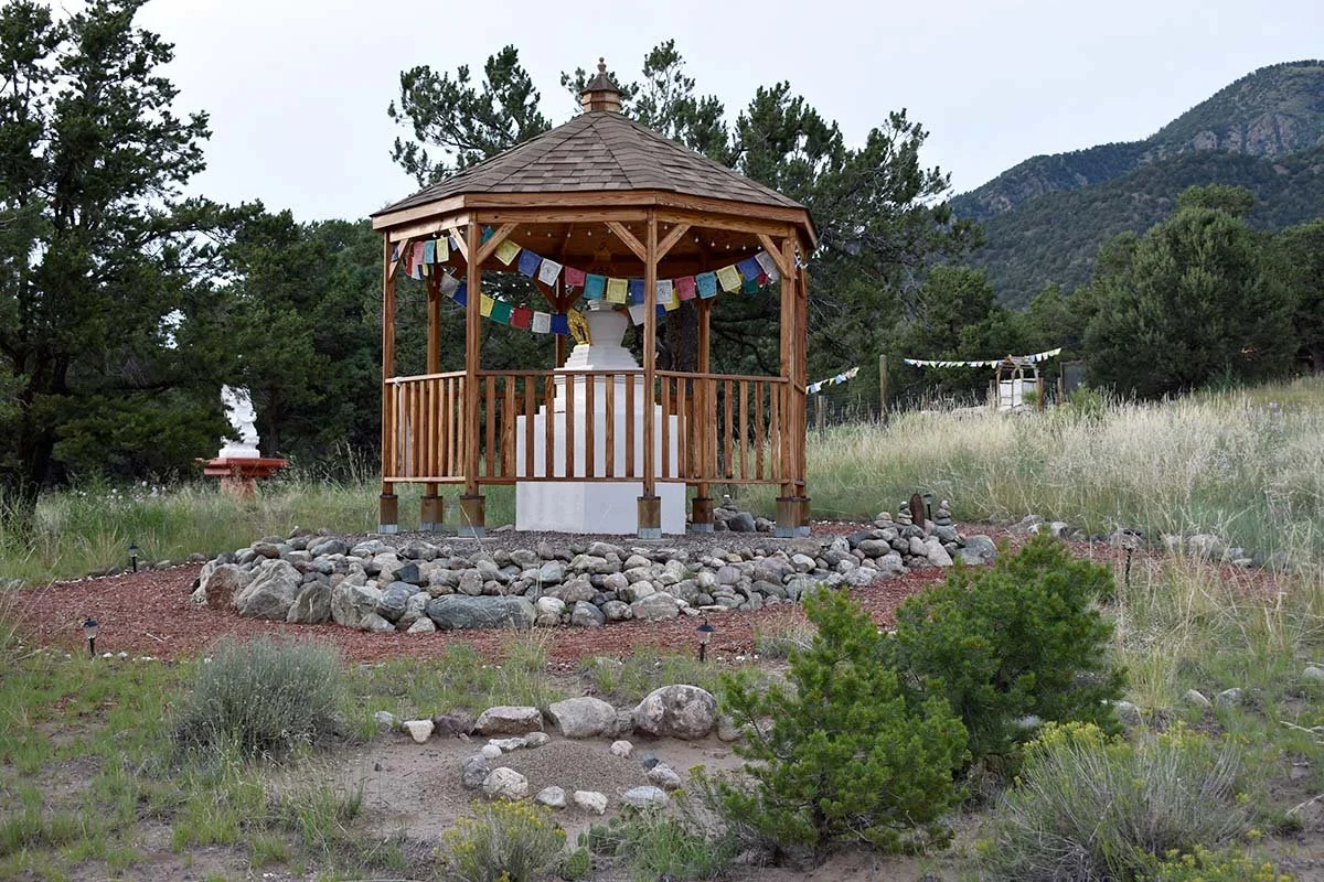 A wooden gazebo with prayer flags surrounds a small white stupa, situated in a grassy outdoor setting with trees, mountains, and a cloudy sky in the background.