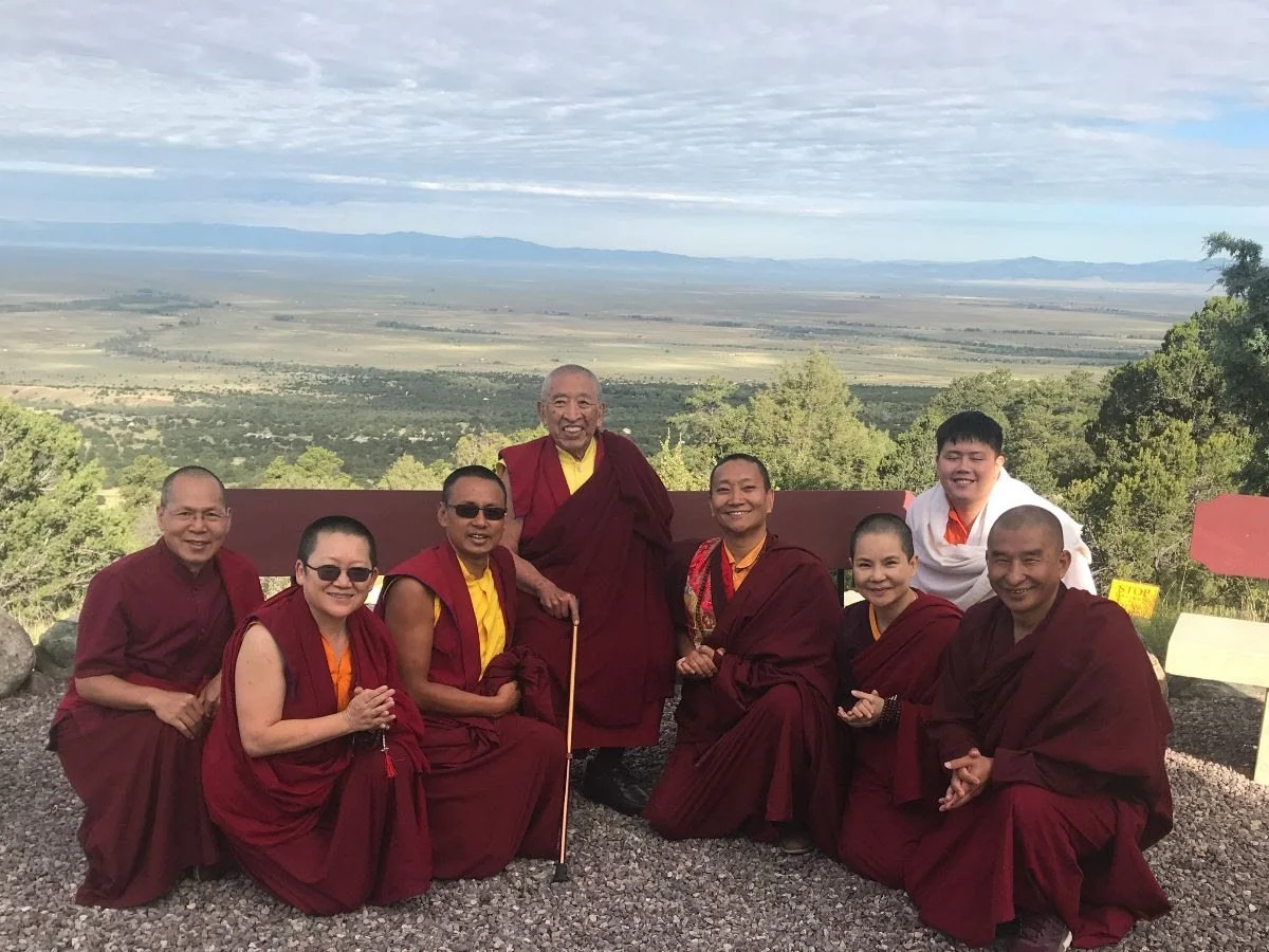 Group of seven people, including monks in maroon robes, smiling outdoors with mountains and trees in the background.