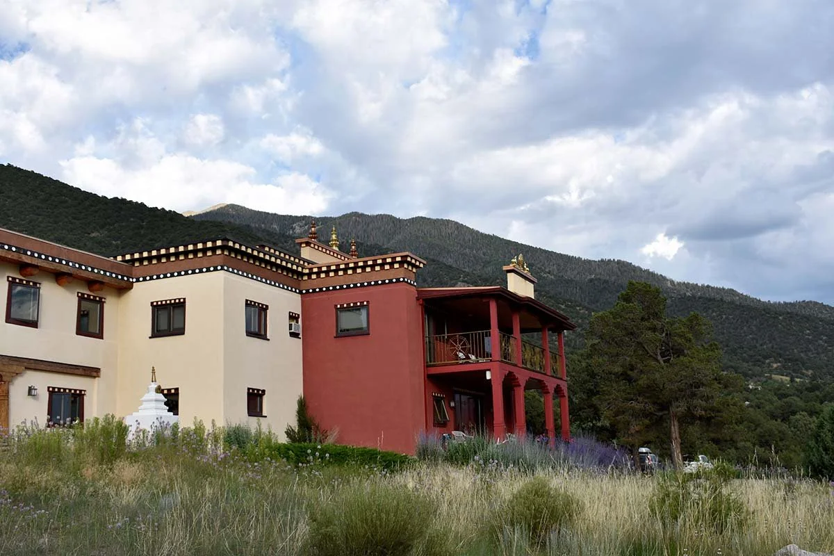 A large, multi-story building with a red and white exterior, surrounded by grassy land and trees, with mountains and a partly cloudy sky in the background.